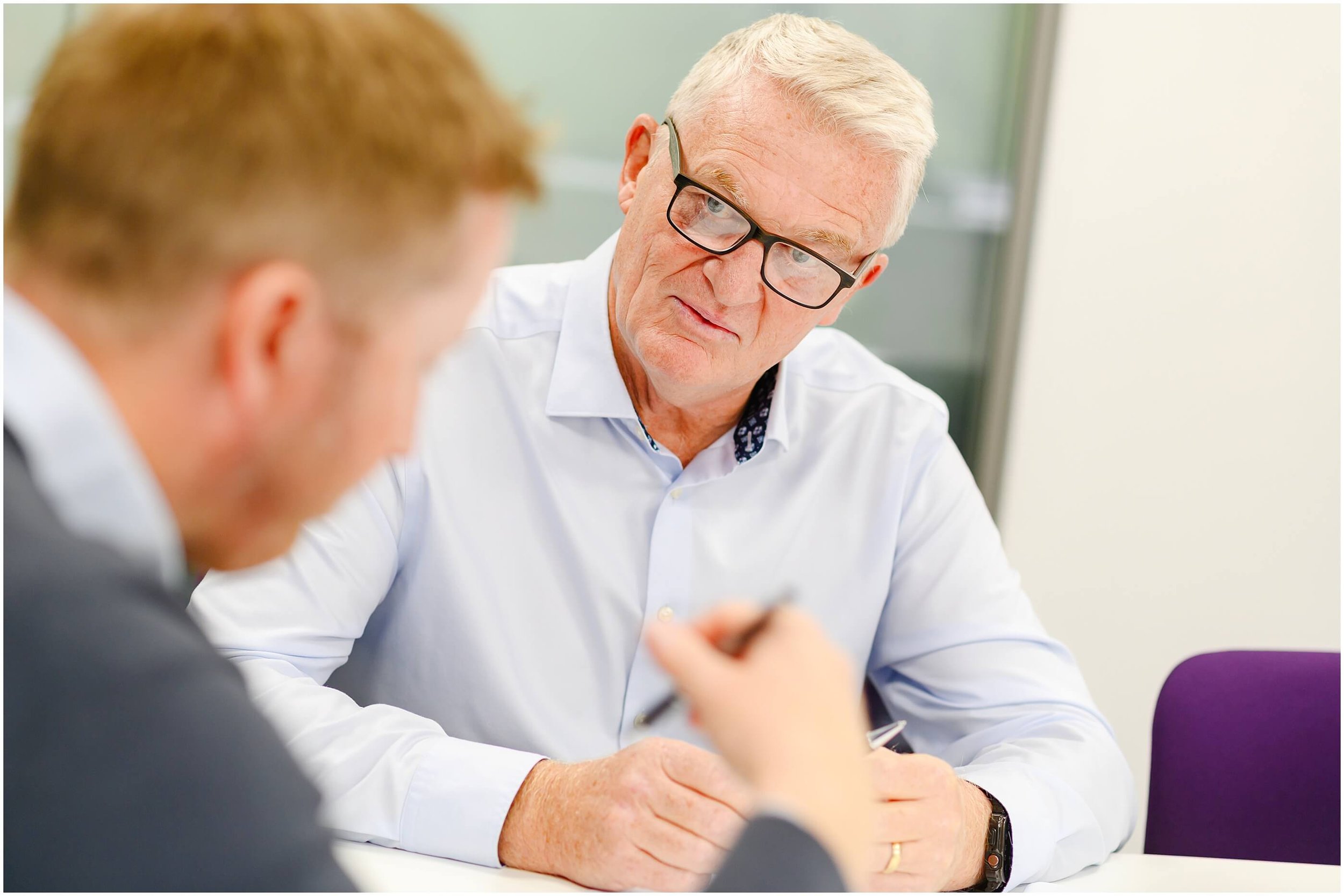 Older man wearing glasses listens during a conversation at a table.