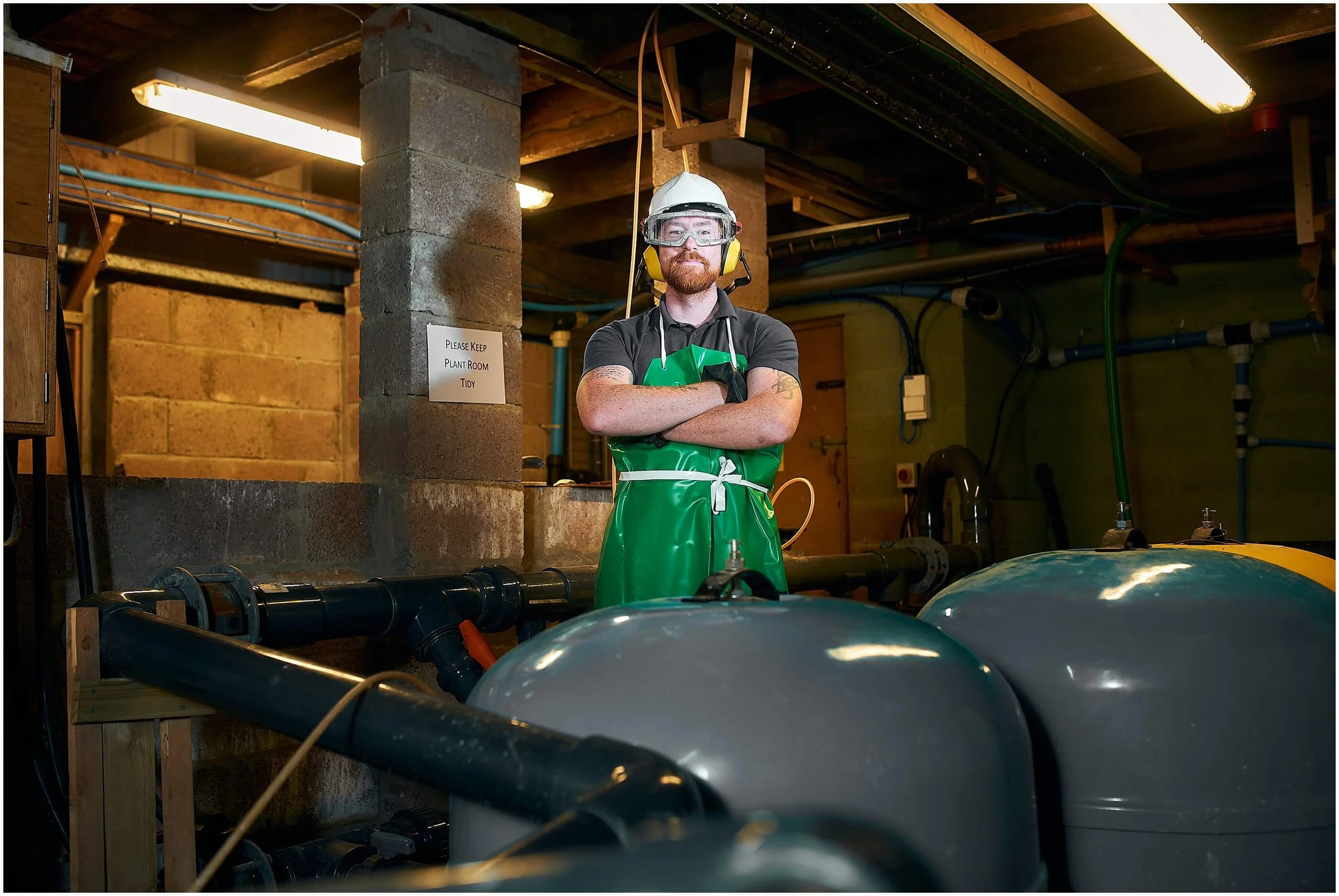Worker wearing a hard hat, safety goggles, hearing protection, and green apron in an industrial plant room with pipes and tanks
