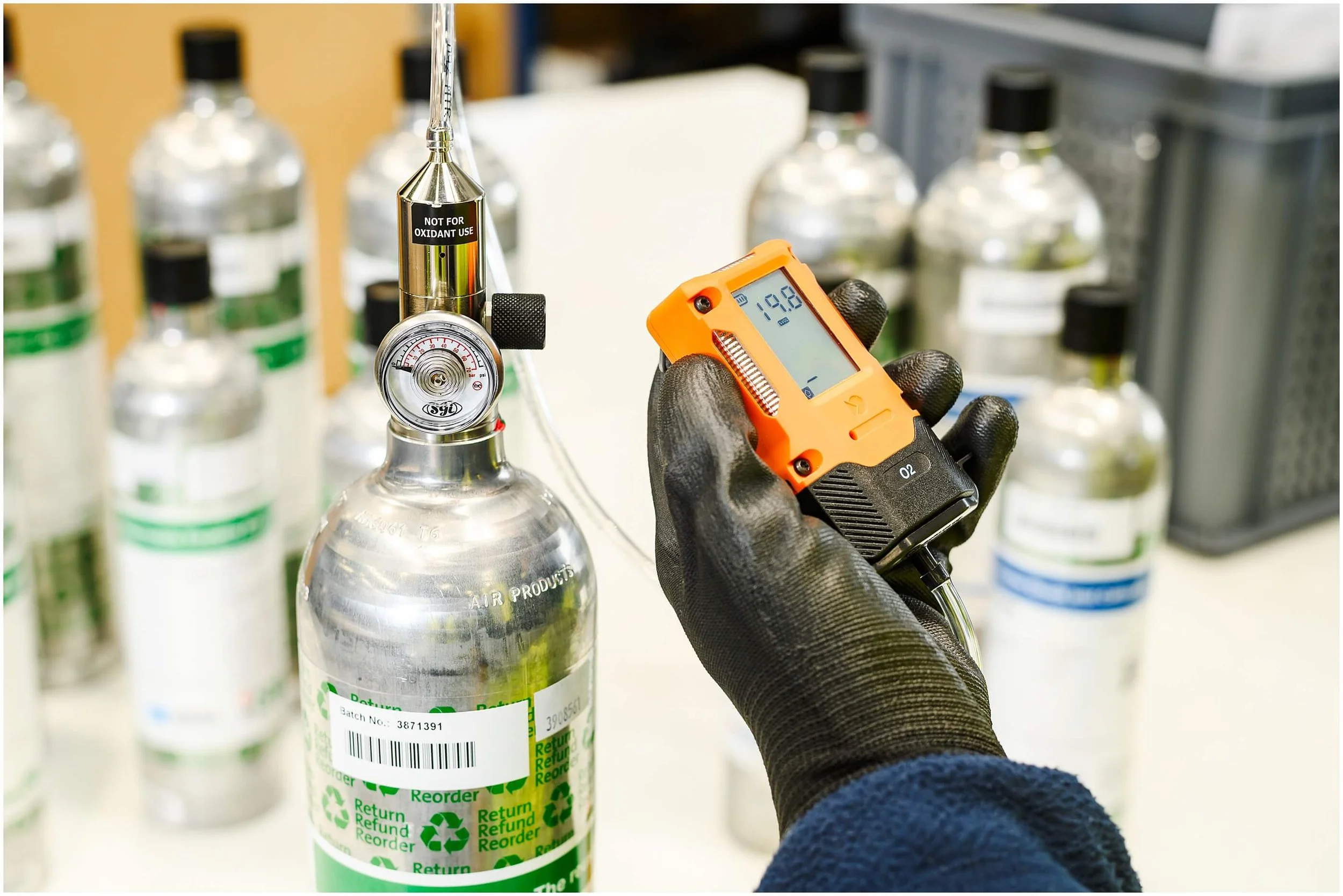 Gloved worker uses an orange oxygen monitor to inspect a medical oxygen cylinder among other gas bottles.