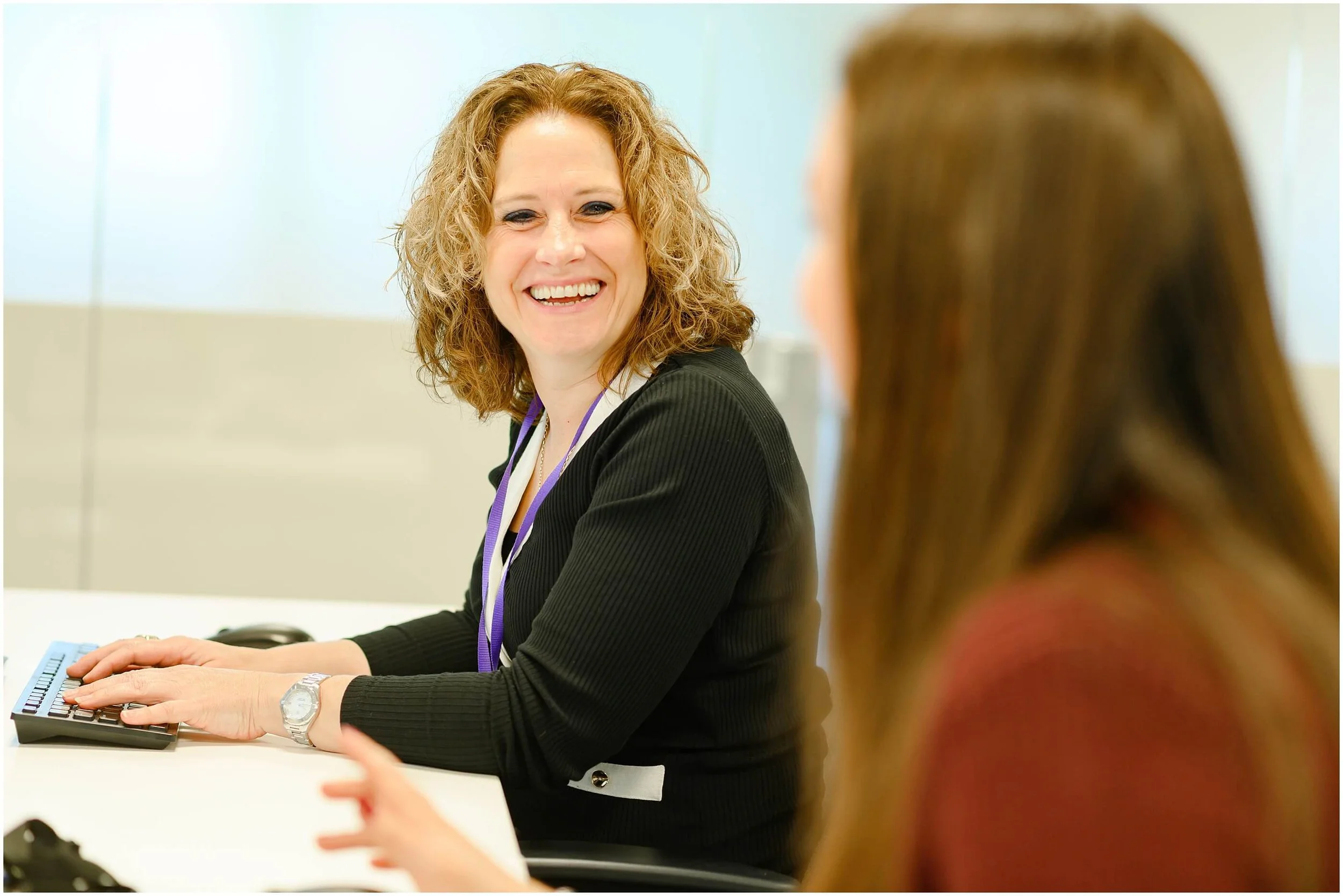 Smiling woman in a professional setting typing on a keyboard while speaking with a colleague