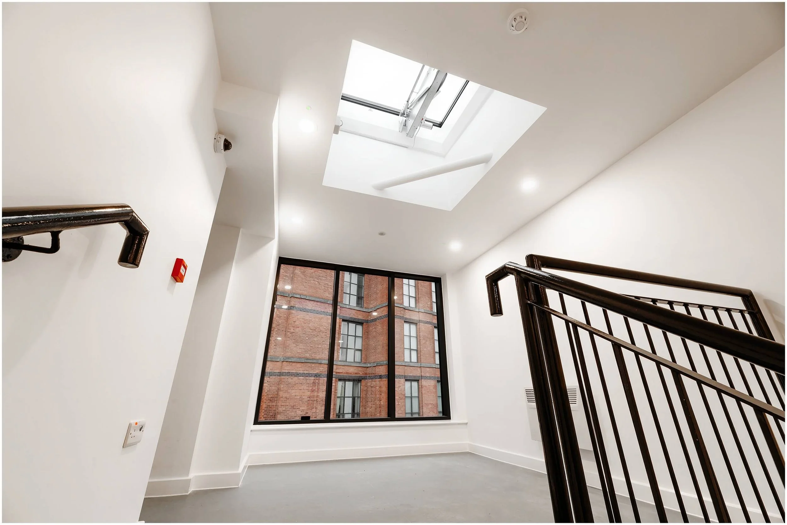 Interior of a modern stairwell with white walls, large window showing a brick building outside, a skylight, and black metal handrails.