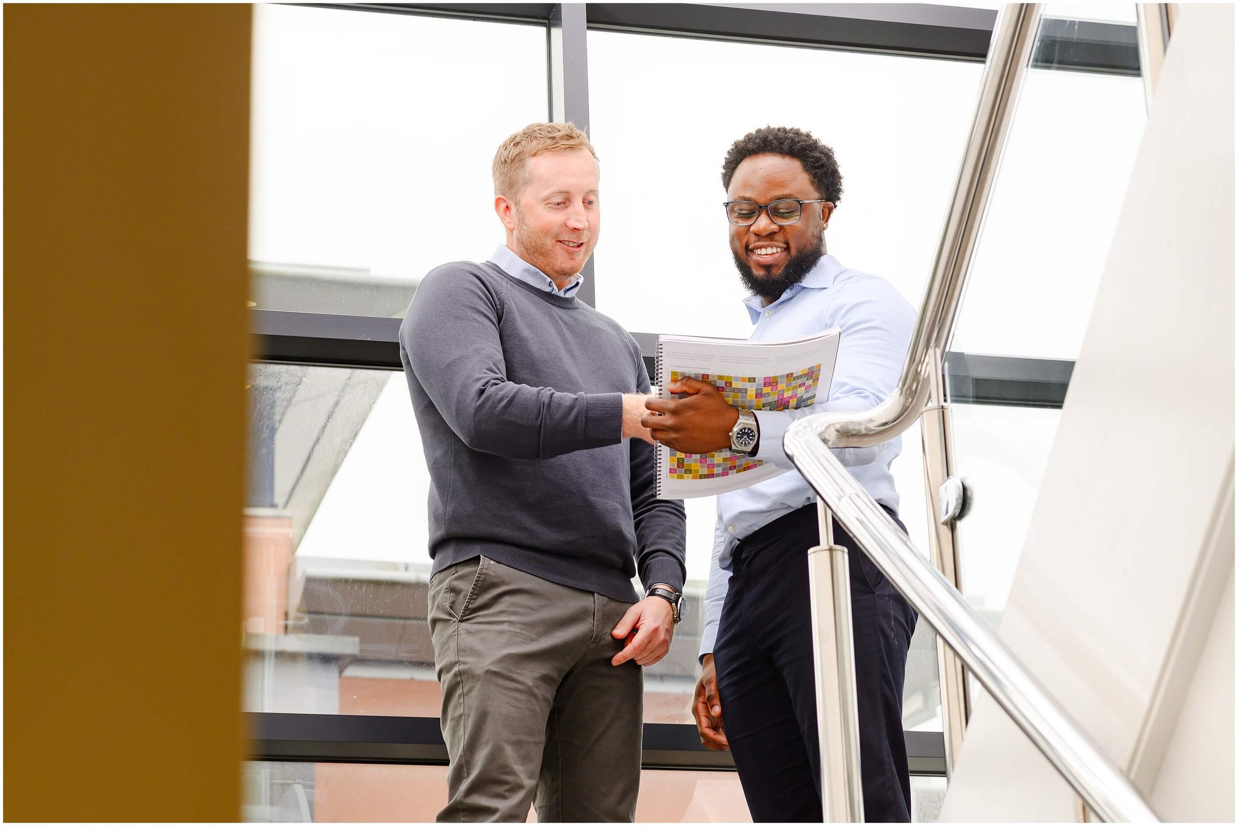Two coworkers discussing a colorful chart while standing on an office staircase.