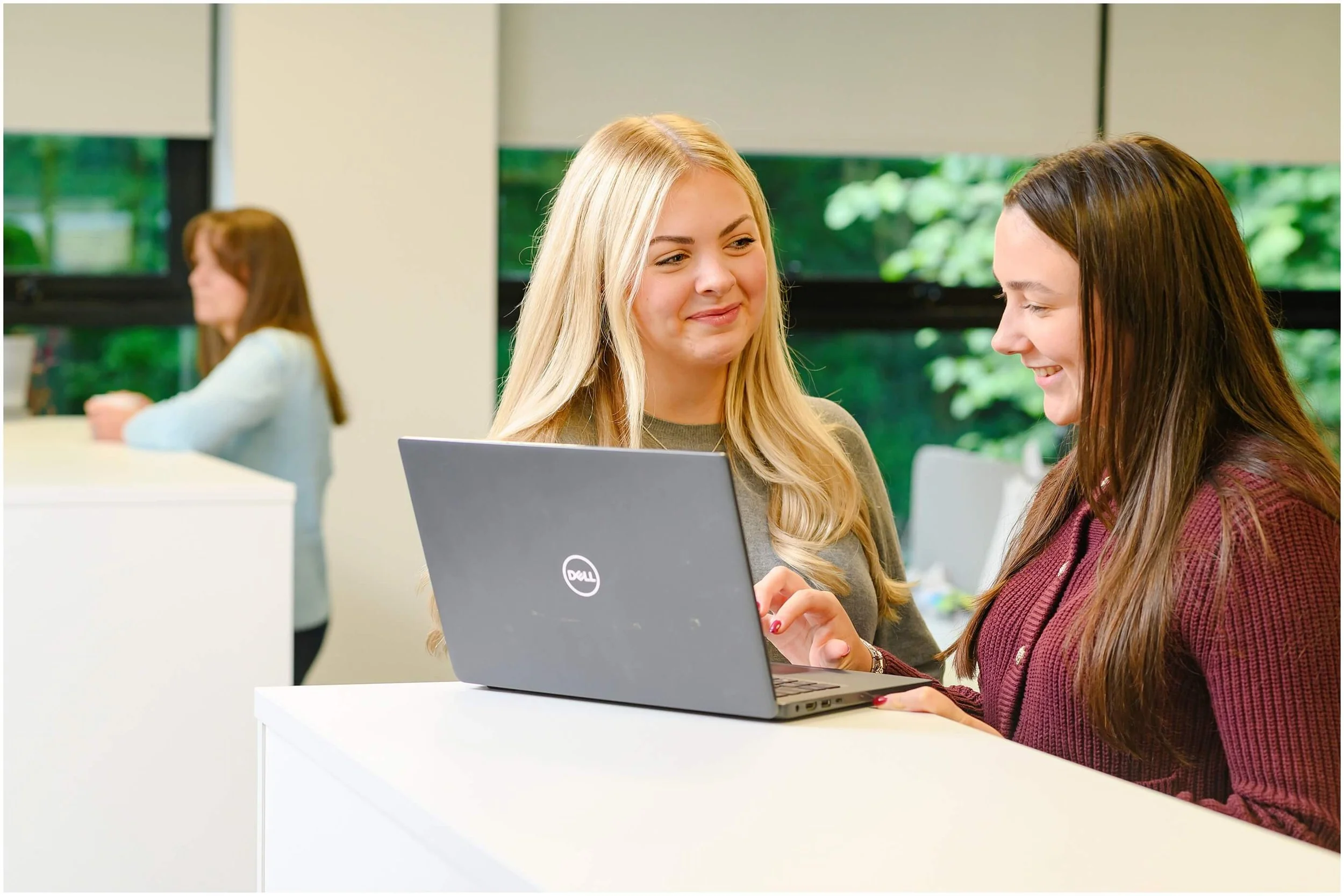 Two women collaborating on a Dell laptop at a table in a bright office, with another person in the background.