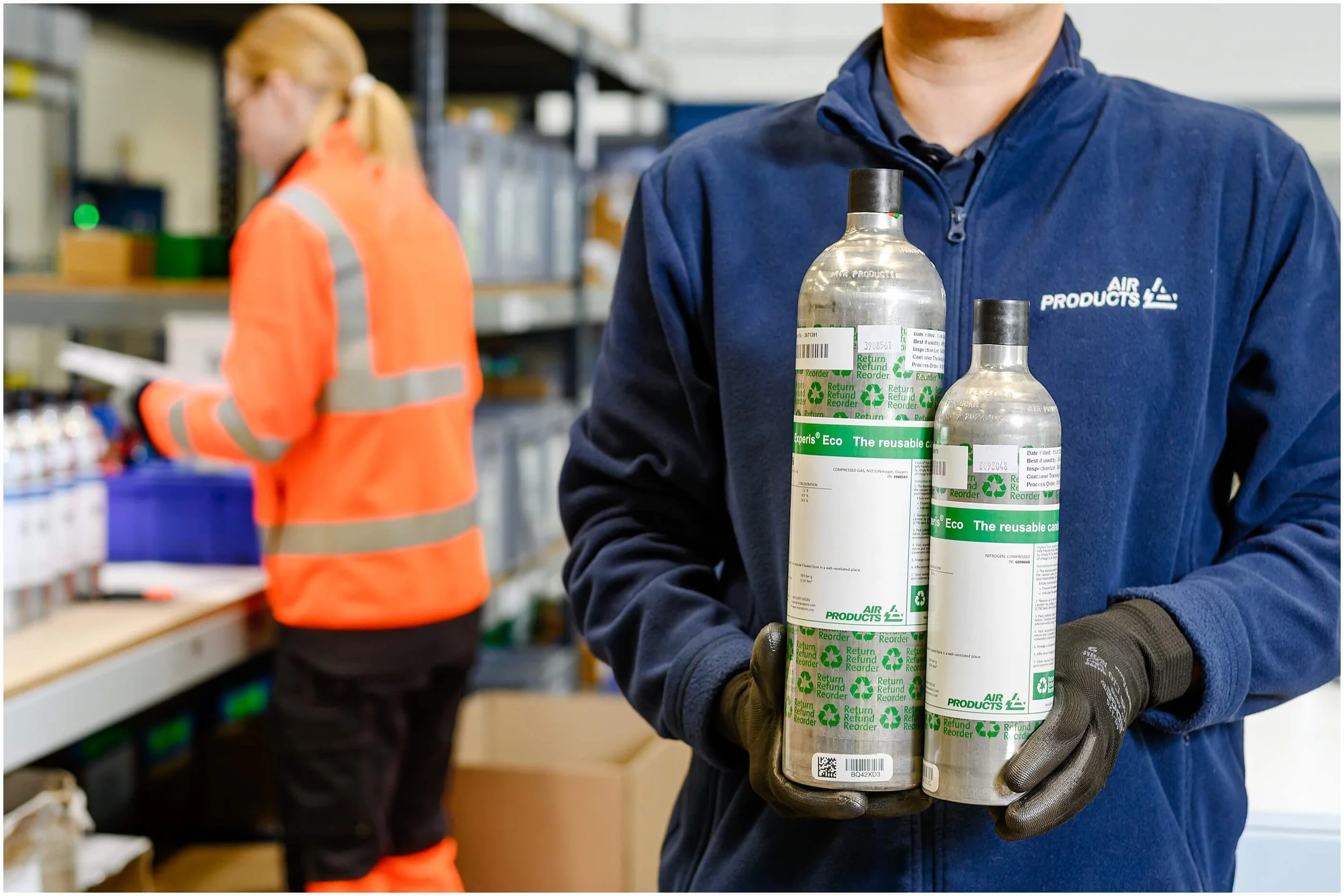 Worker in a blue jacket and gloves holding two reusable compressed gas cylinders in an industrial facility