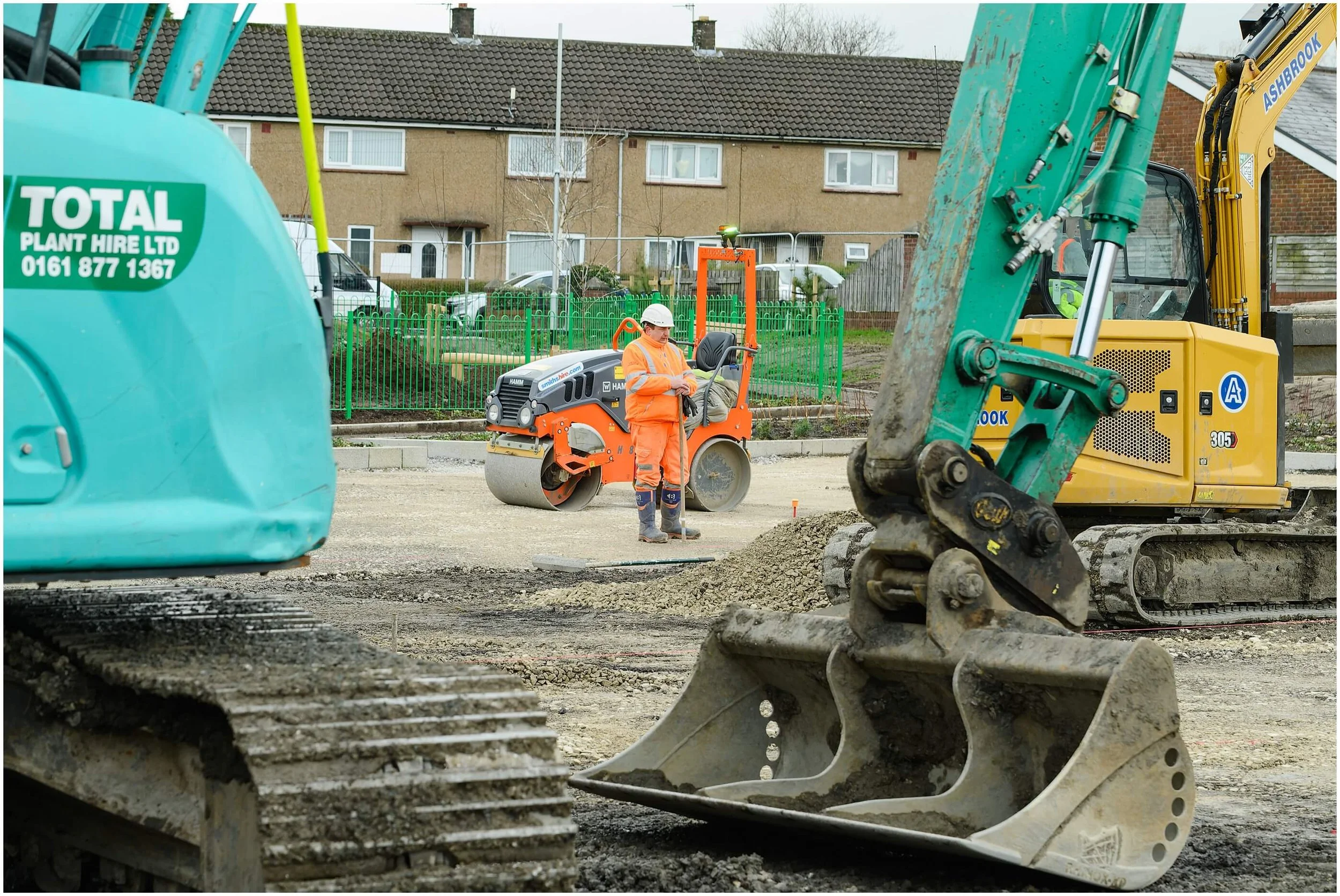 Construction worker in high-visibility clothing beside a road roller and excavators at a residential road construction site