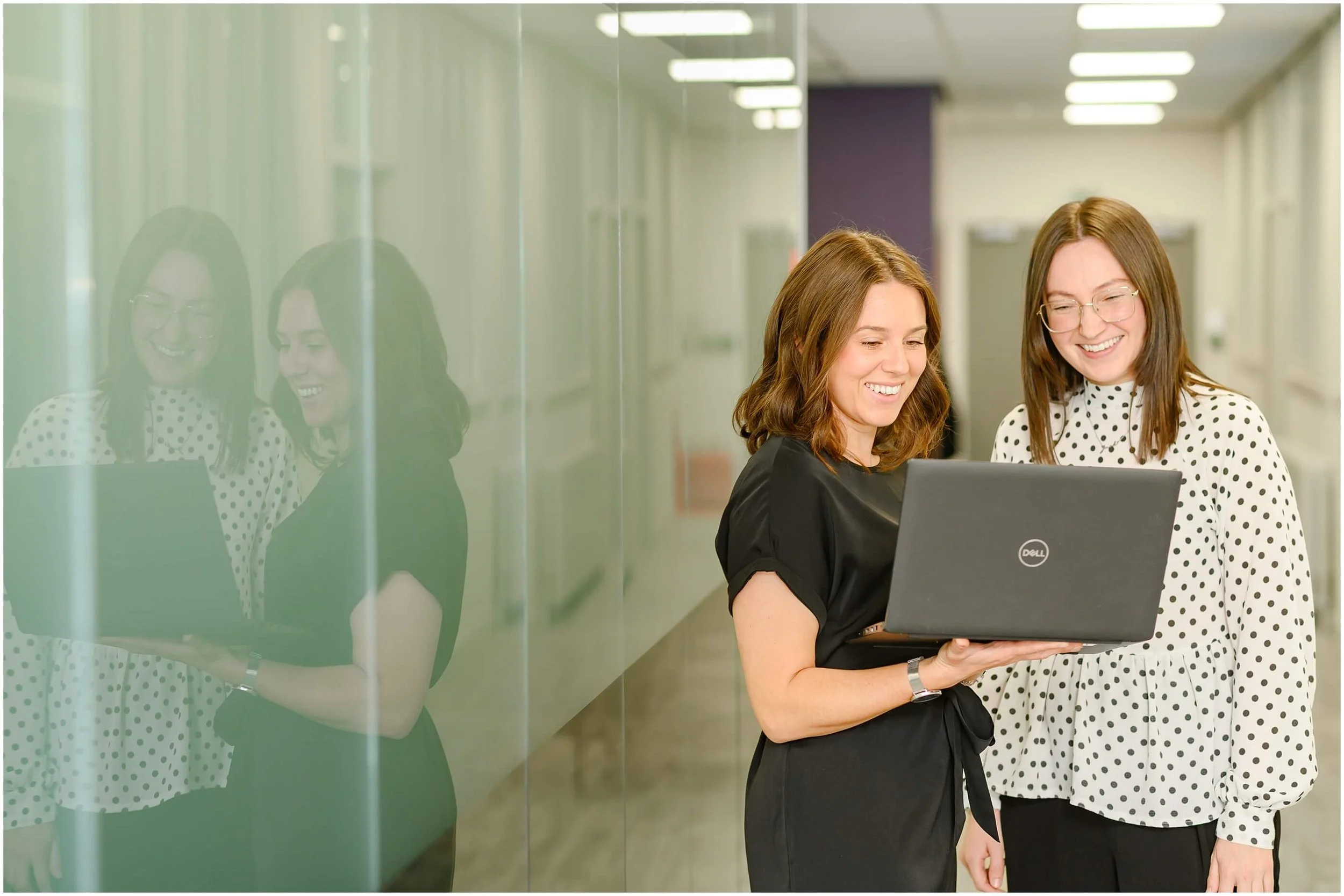 Two smiling businesswomen reviewing a Dell laptop in a modern office hallway