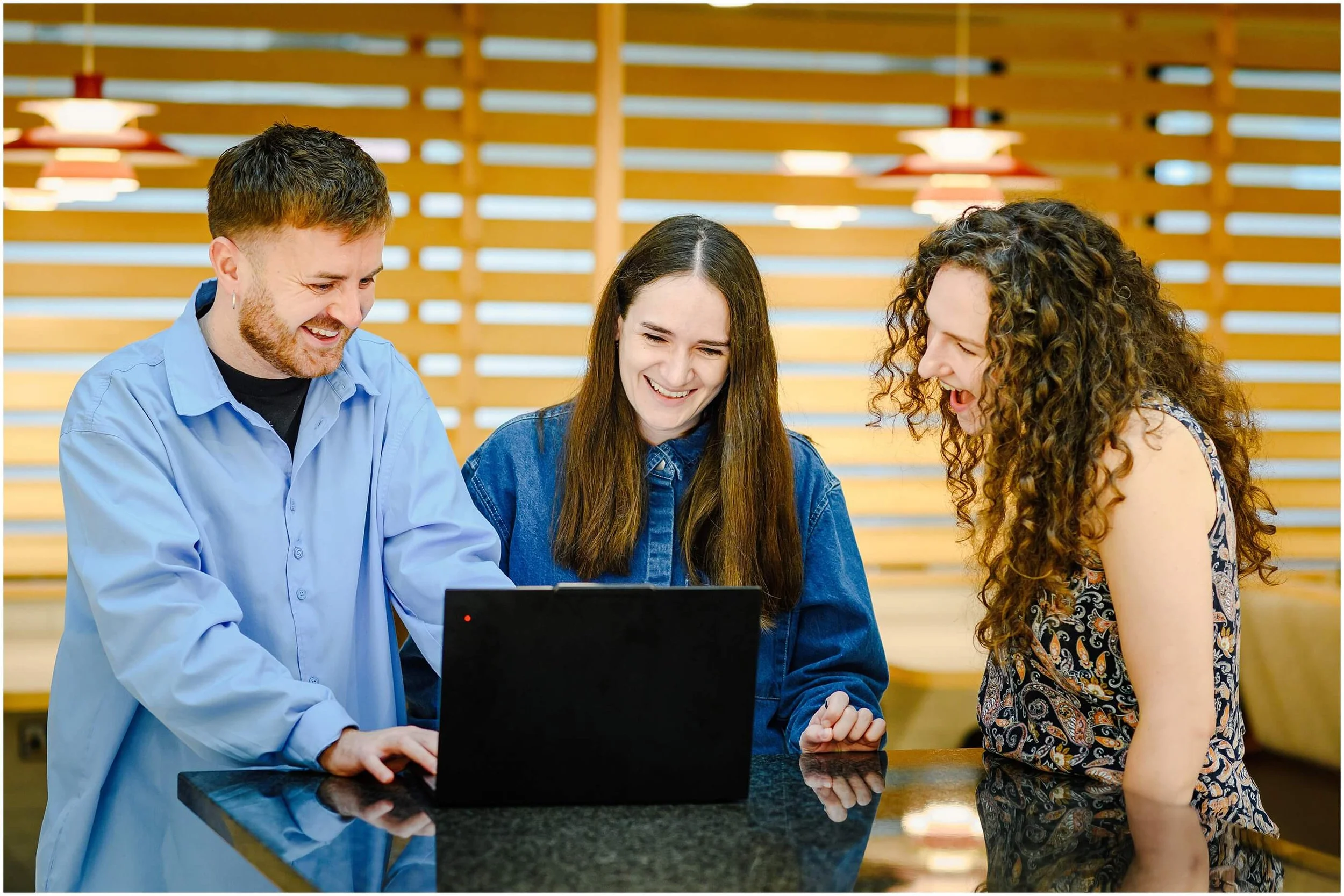 Three coworkers laughing together around a laptop in a bright café