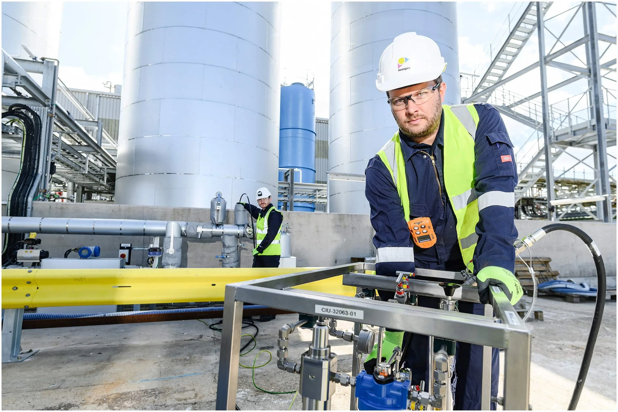 Industrial technicians in hard hats and high-visibility vests working beside storage tanks, pipes, and processing equipment at a chemical facility