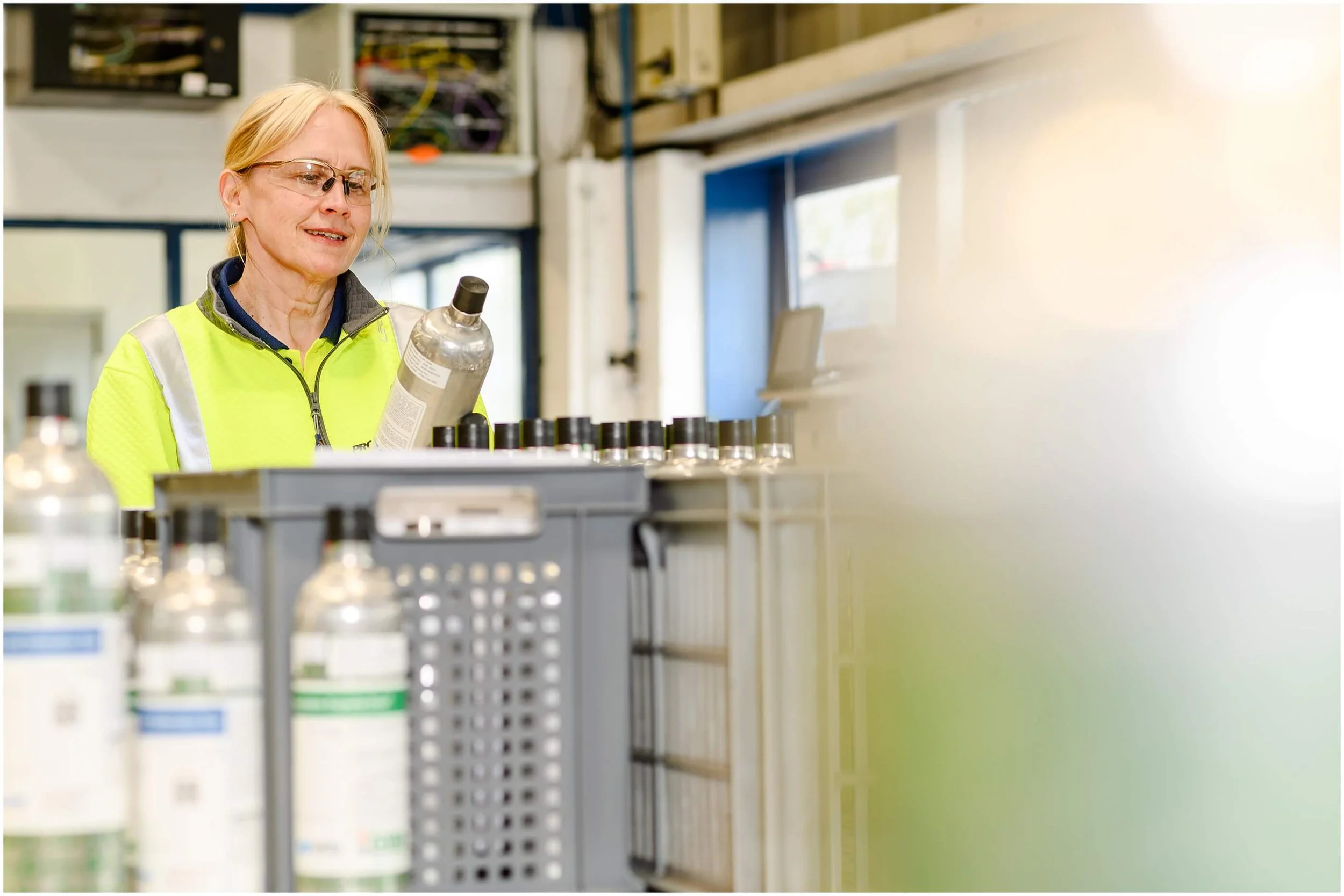 Factory worker in a high-visibility jacket inspecting a metal container among industrial bottles and equipment