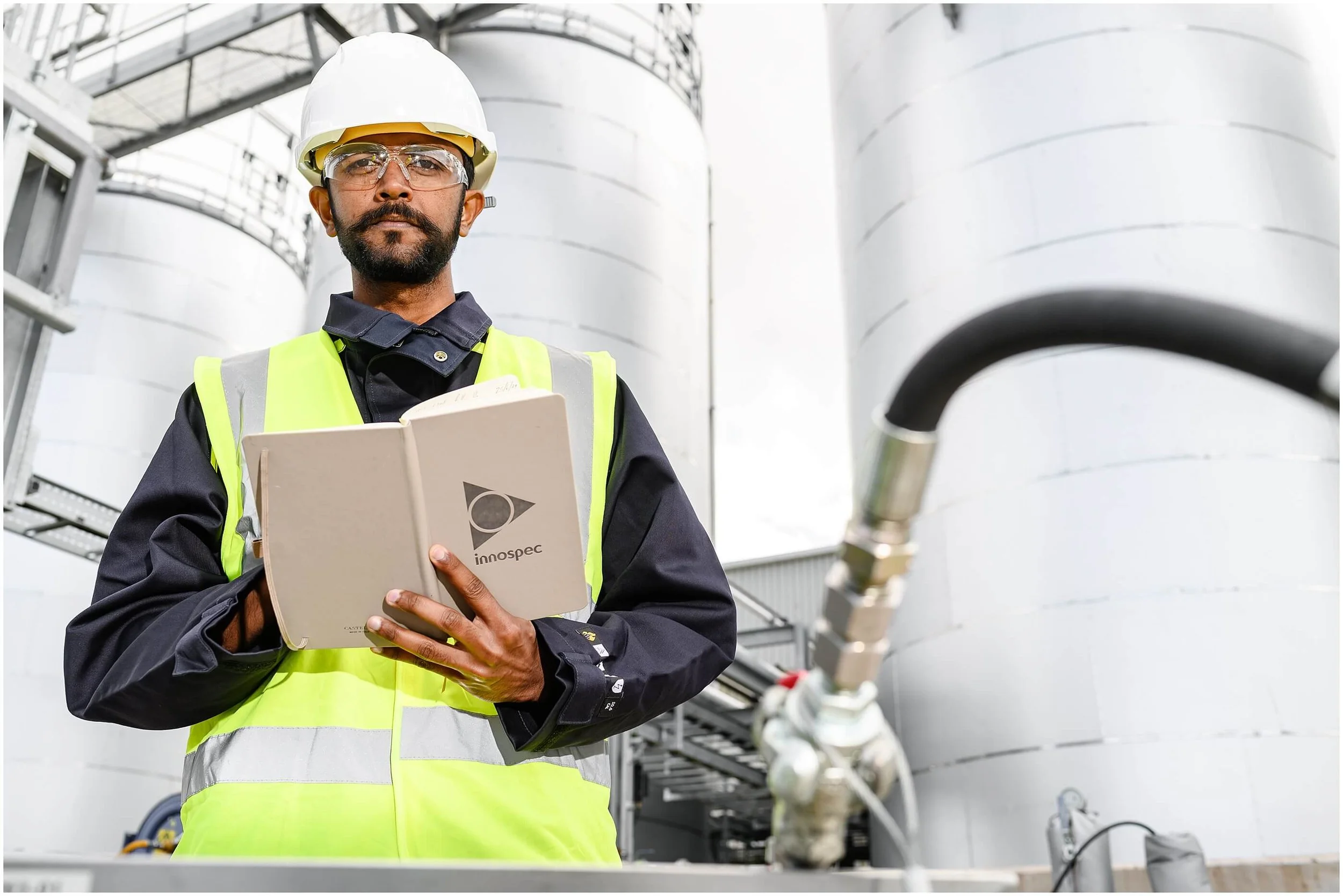 Industrial worker wearing a hard hat and safety vest, holding a clipboard in front of large storage tanks