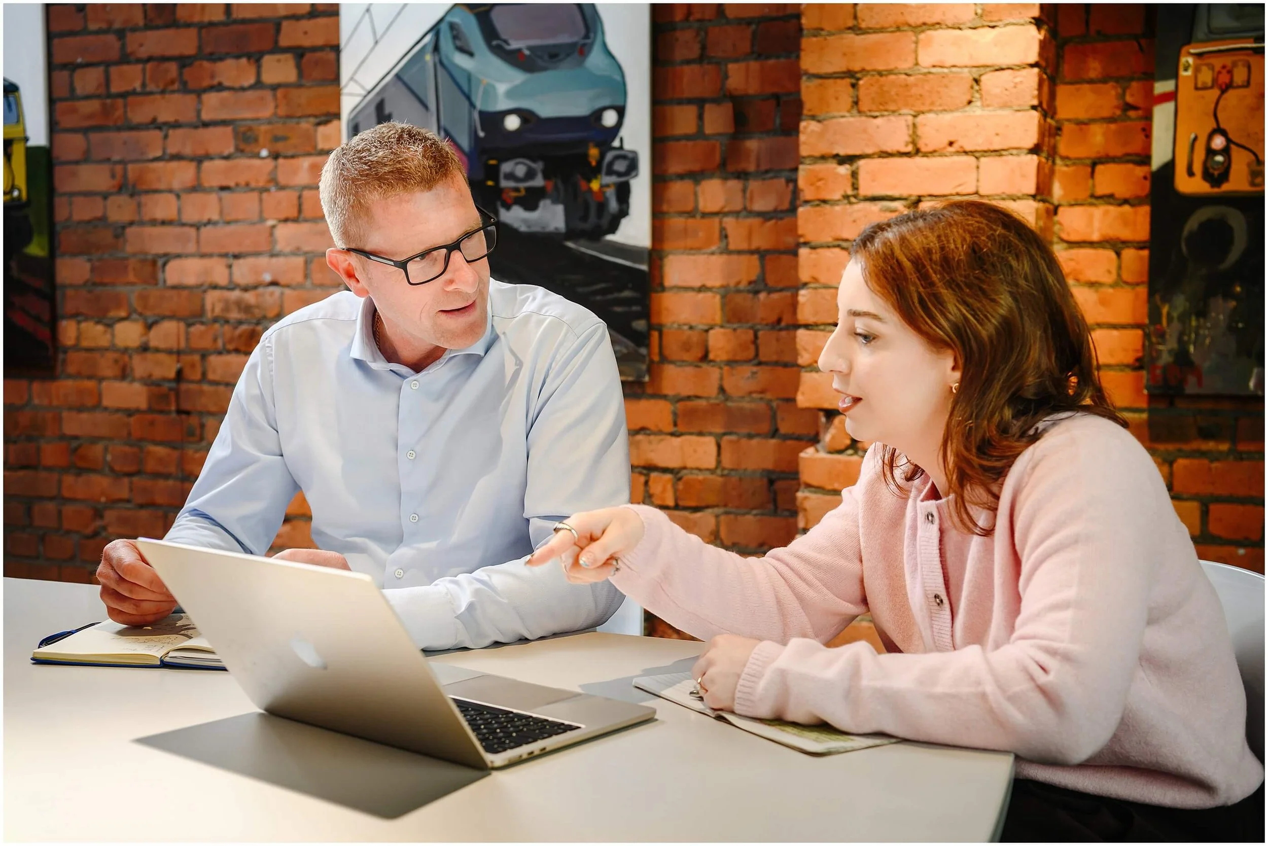 Two coworkers discussing a project over a laptop at a table in a brick-walled office