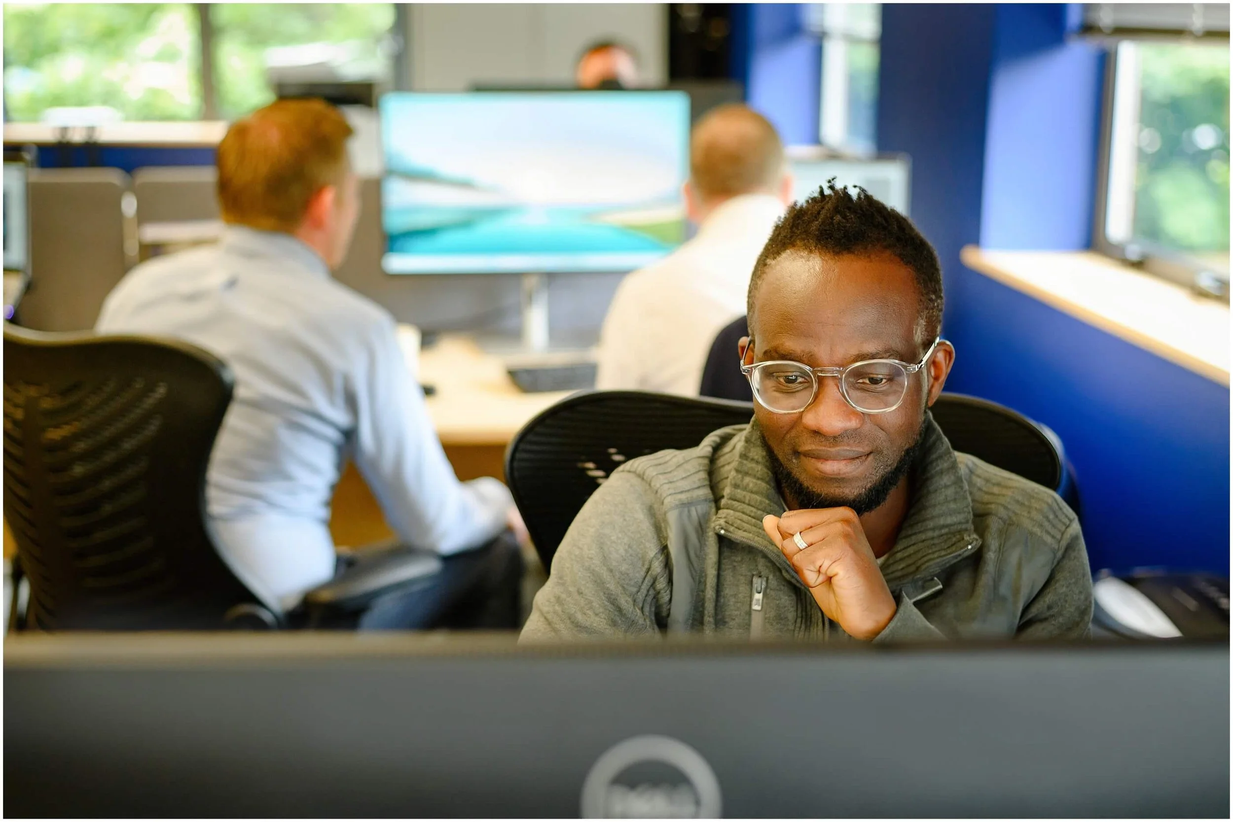 Man wearing glasses working at a computer in a bright modern office, with coworkers and monitors in the background.