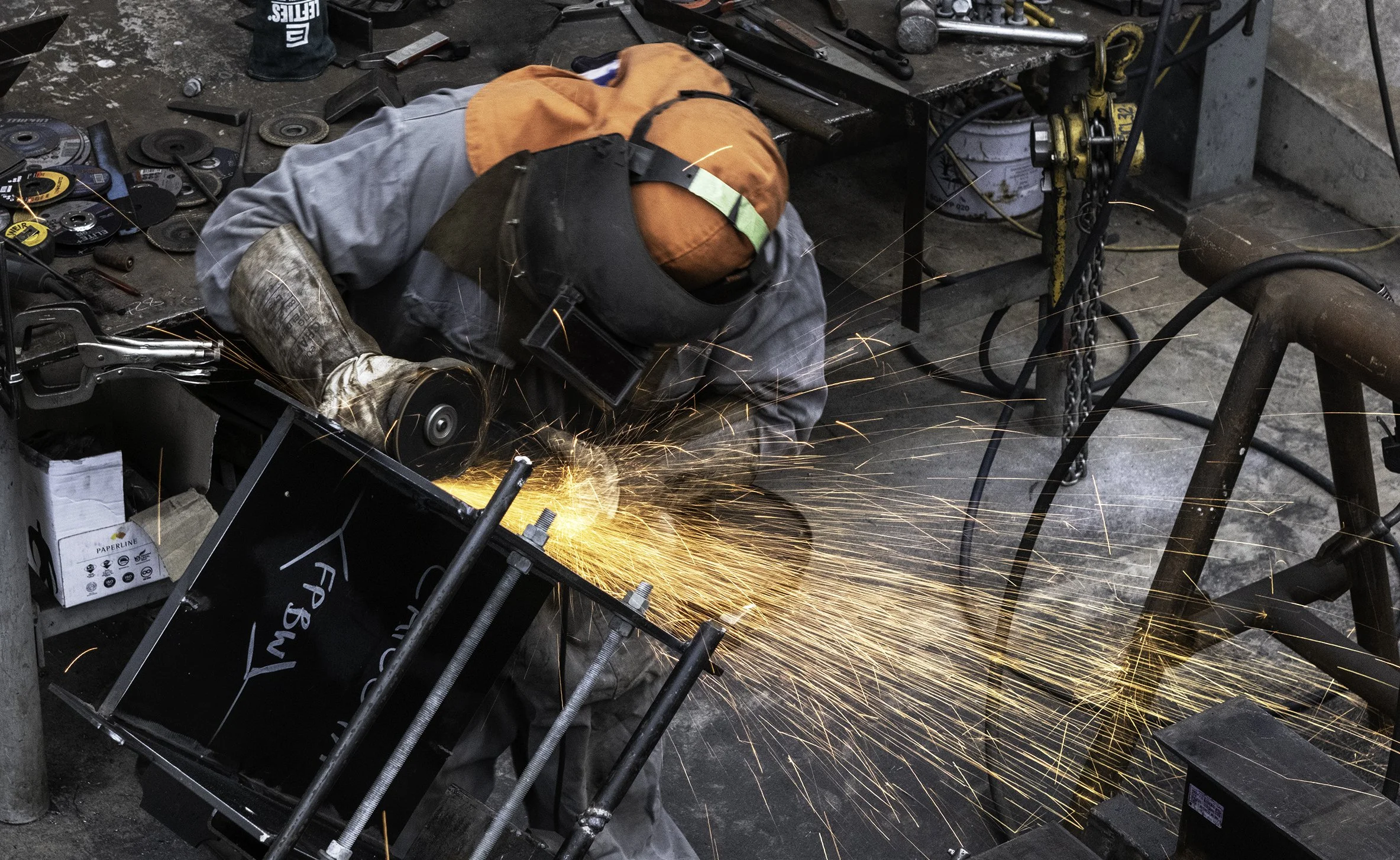 A worker wearing safety goggles, a vest, gloves, and an orange hood is welding metal, with bright sparks flying out. The workshop is at Warner Constructions fabrication and engineering New Plymouth.