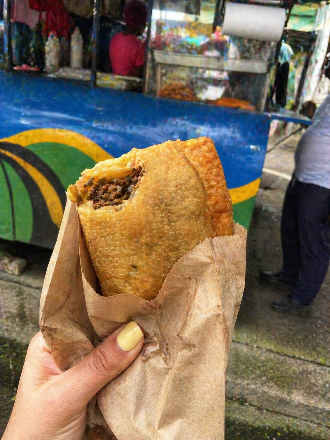 A hand holding a freshly fried Pati beef turnover wrapped in paper bag at a street food stall in Puerto Limón, Costa Rica.