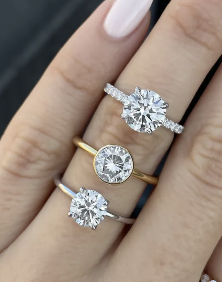 Close-up of three fingers with different engagement rings featuring large round-cut diamonds, on a person's hand with light skin and painted nails.