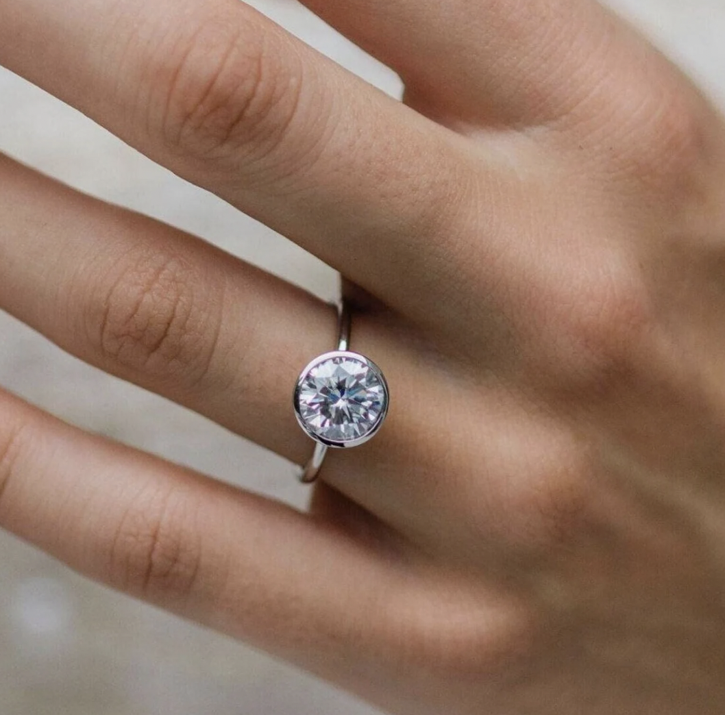 A hand with a silver ring featuring a large round diamond on the ring finger.