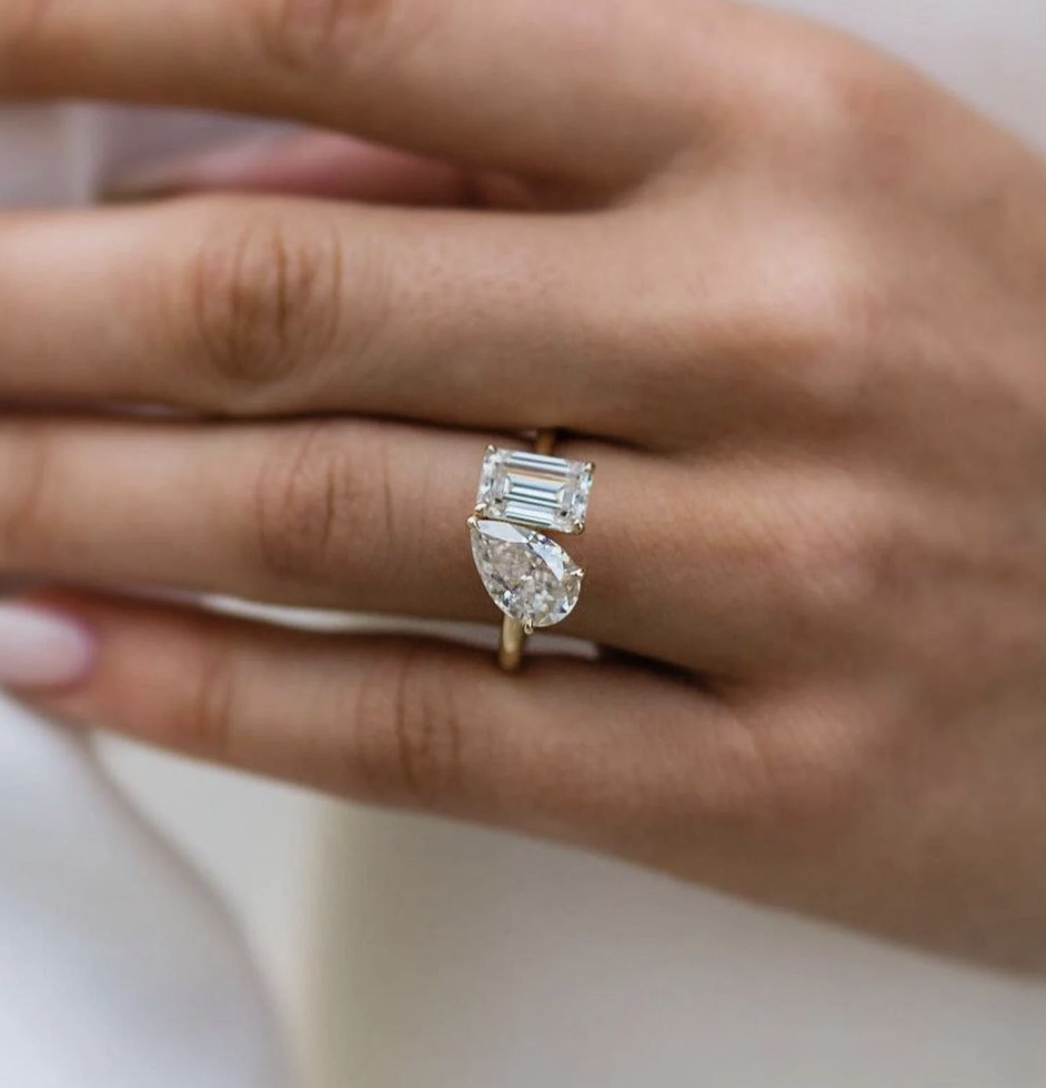 Close-up of a woman's hand wearing a gold ring with a large rectangular emerald-cut diamond and a pear-shaped diamond set on a simple gold band, against a light background.