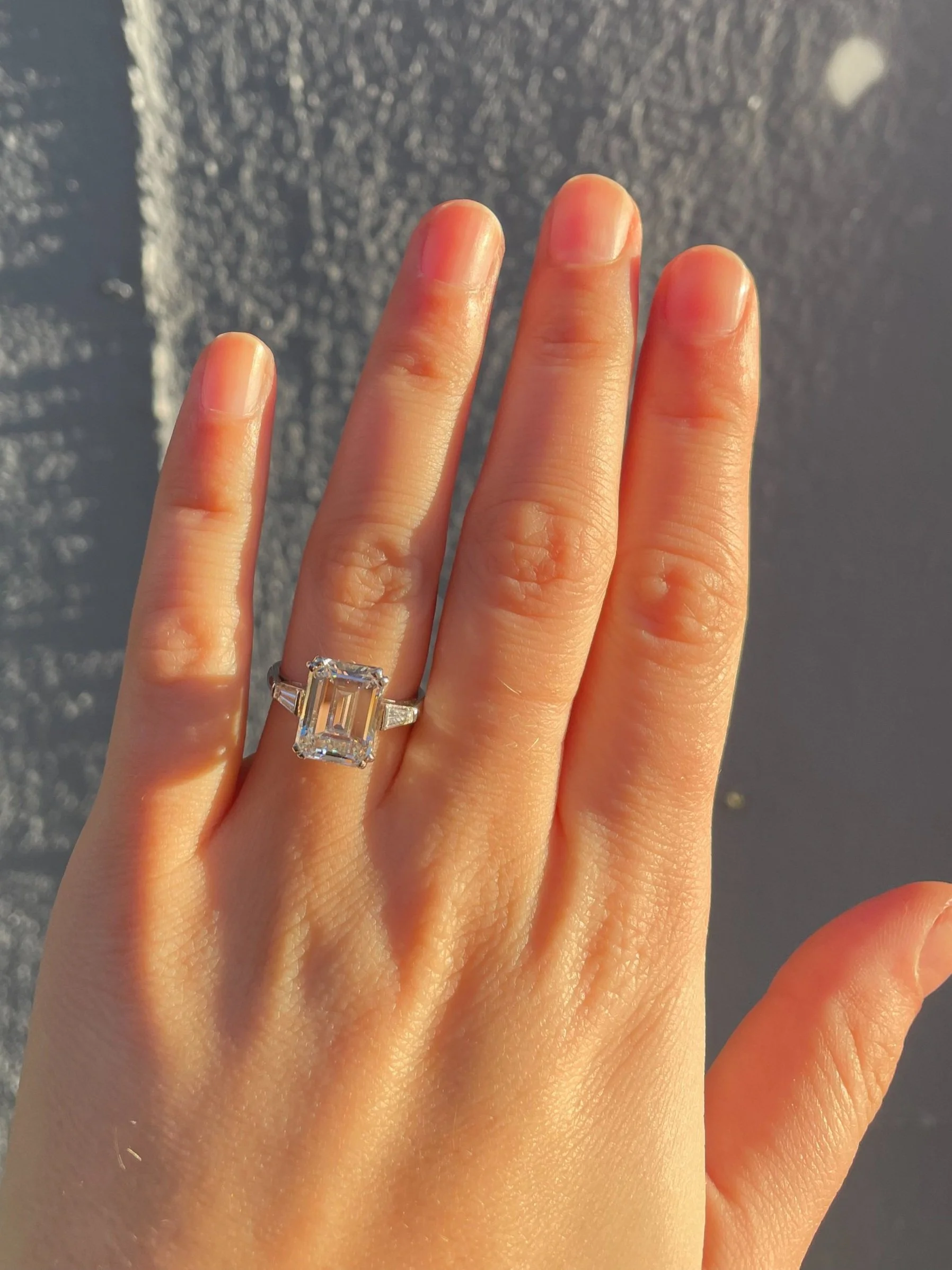 A hand with a large emerald-cut diamond ring on the ring finger, shown against a textured gray background.