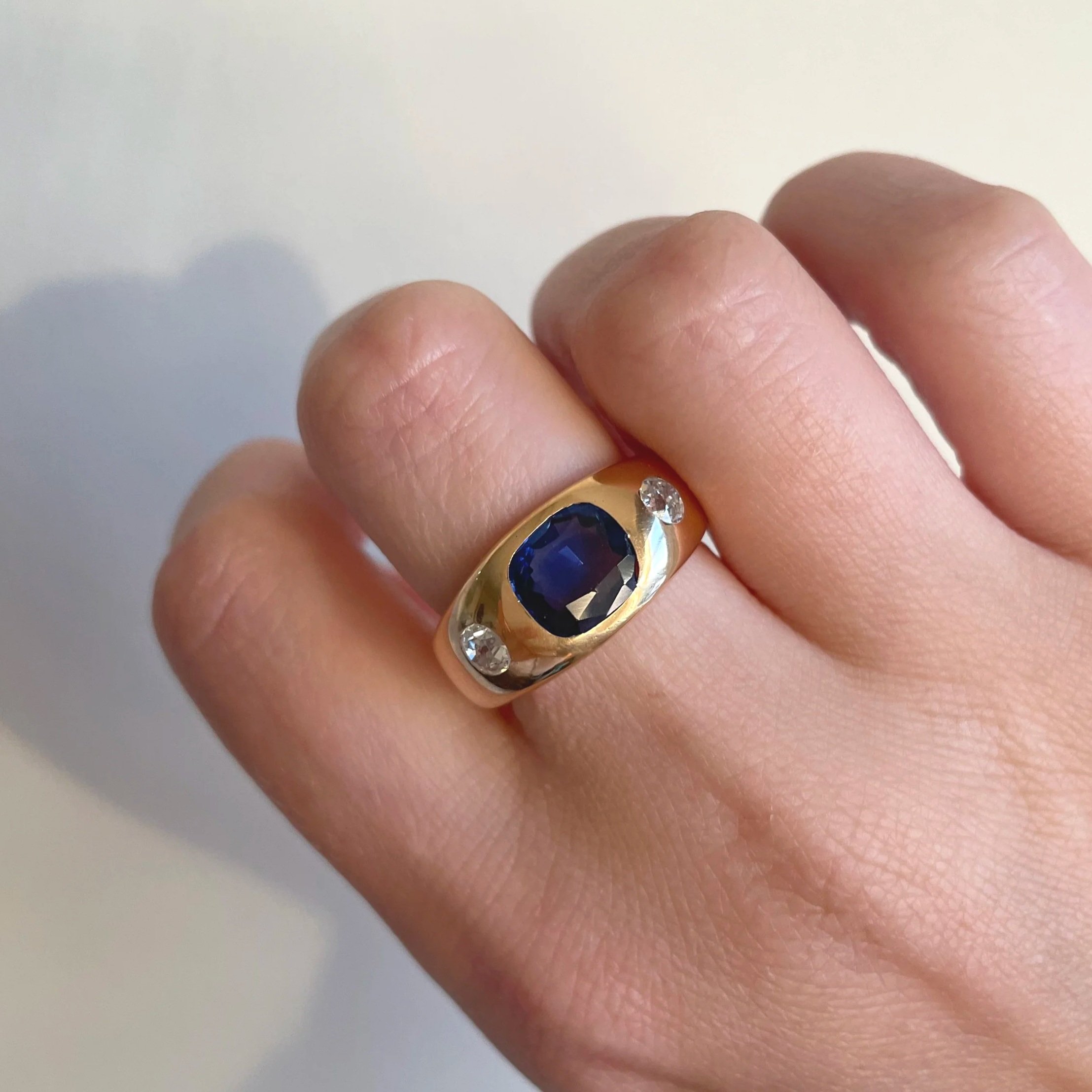 Close-up of a person's hand wearing a gold ring with a large dark blue gemstone and small clear gemstones.