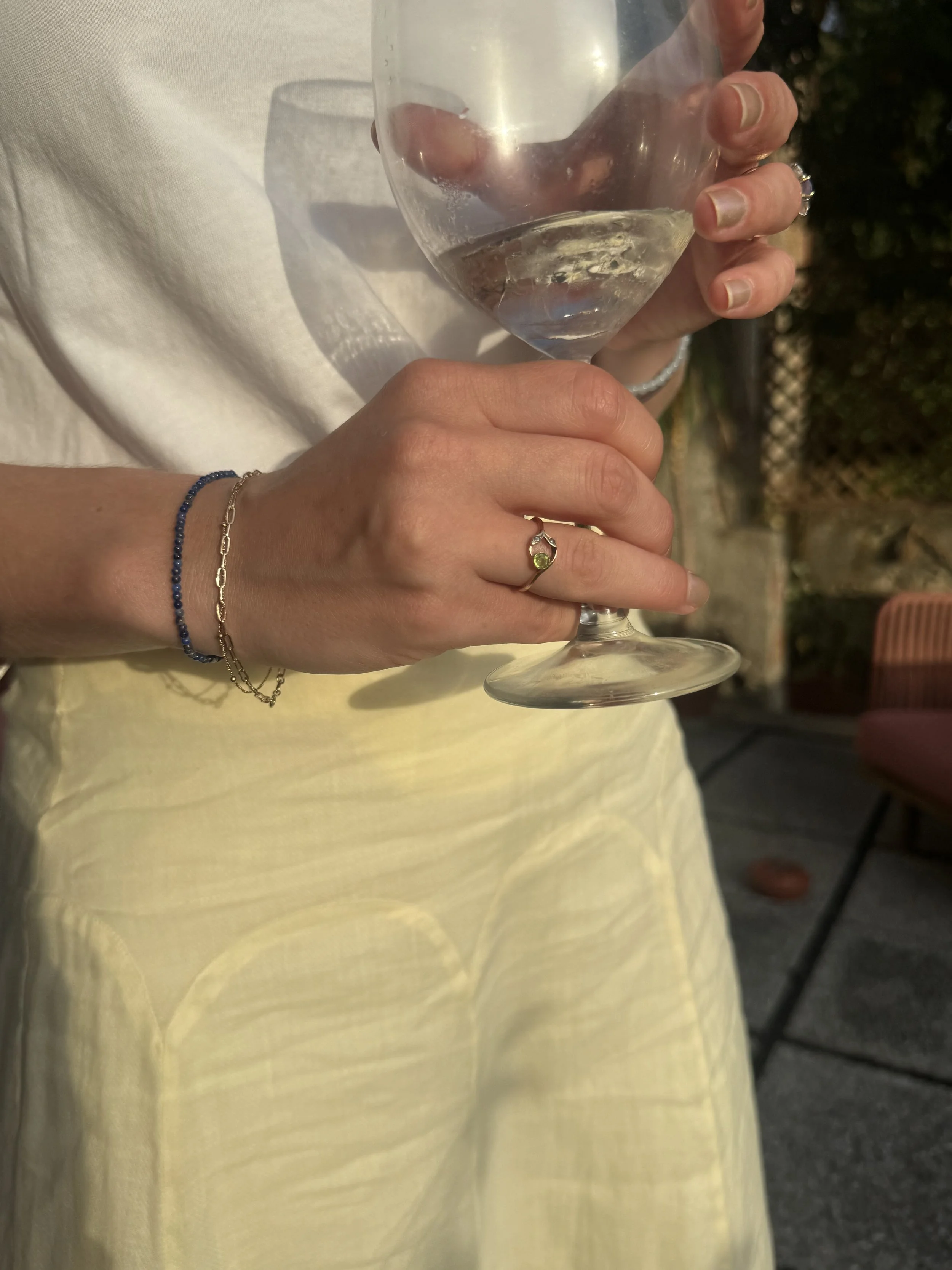 A person with a ring on their middle finger, wearing a yellow dress, holding a transparent wine glass with light-colored wine inside. The person's right wrist has a blue beaded bracelet and a gold chain bracelet. The background shows outdoor evening or night setting with chairs and fence.