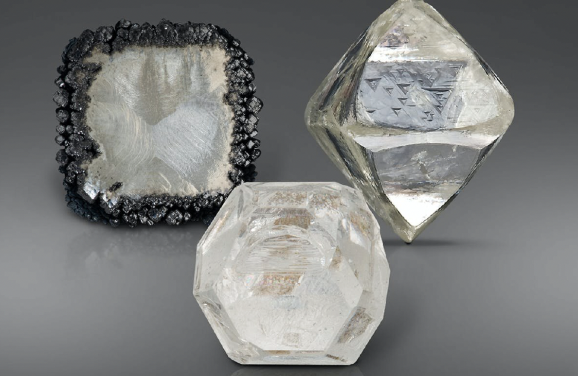 Three large, faceted quartz or crystal objects with geometric shapes and reflective surfaces, arranged on a gray background. One has a rough, bubble-like black border and the others are smooth and transparent with different polygonal shapes.