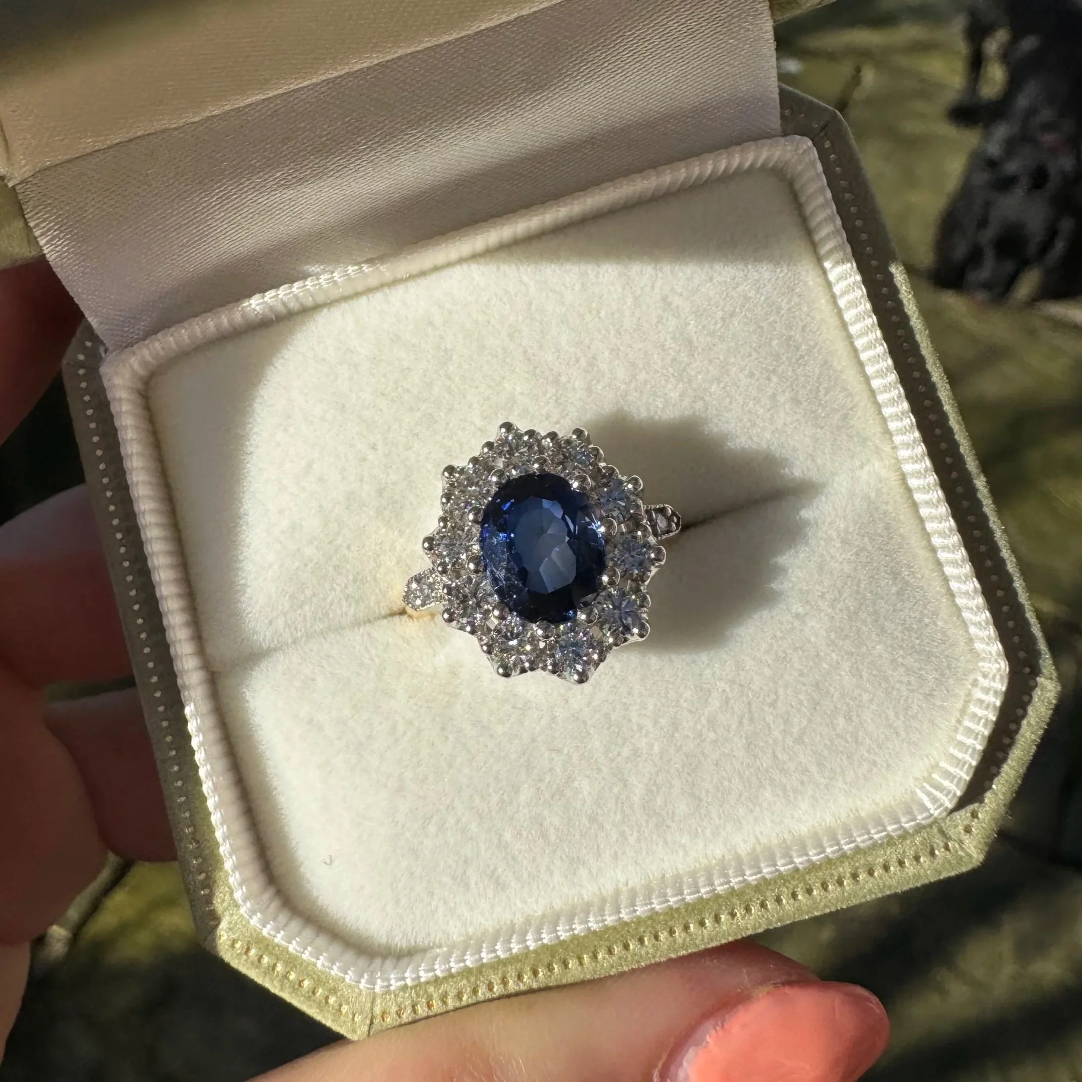 A close-up of a ring with a large blue gemstone surrounded by smaller clear stones, placed inside a cream-colored jewelry box with a satin lining.