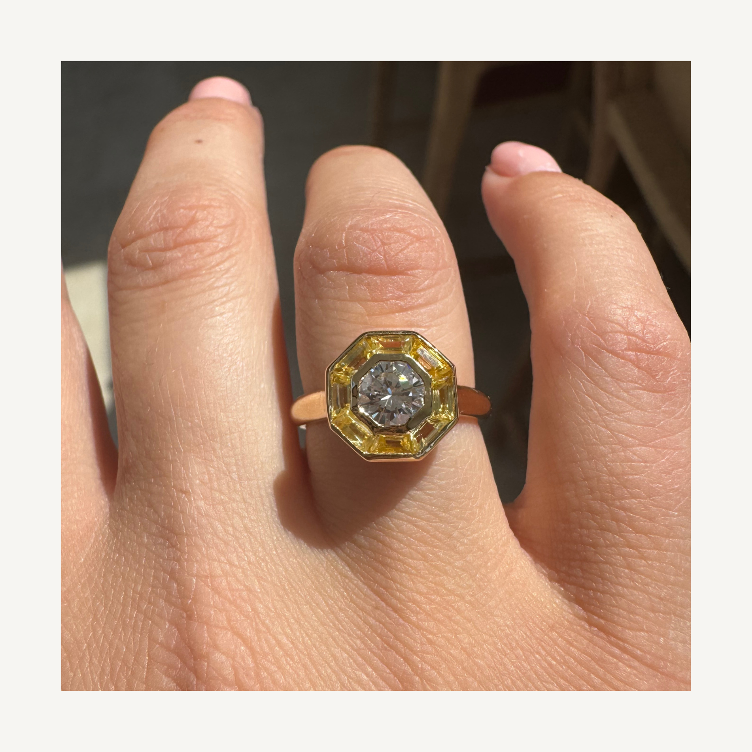 A hand wearing a ring with a central diamond surrounded by yellow rectangular stones, set in gold.