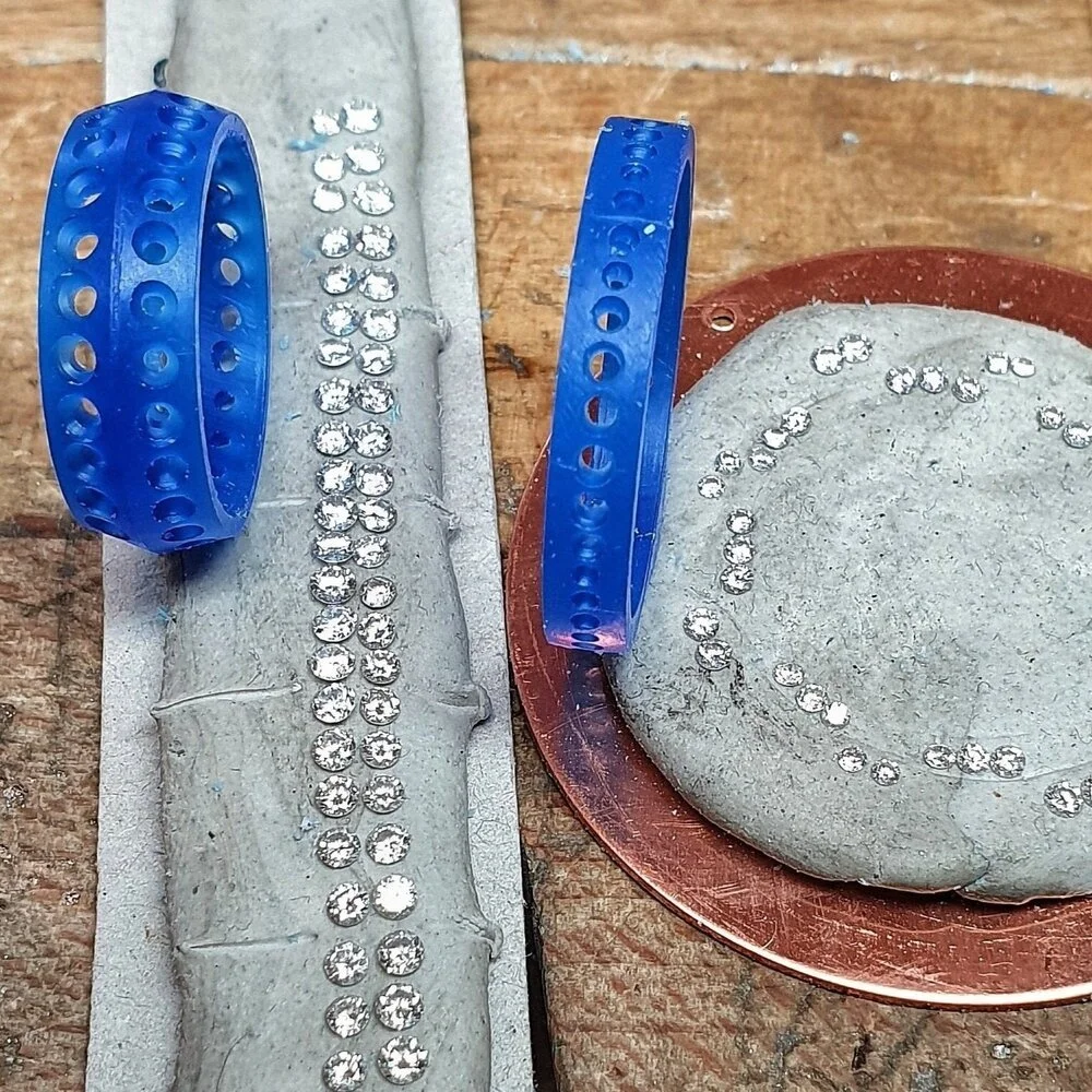 A diamond ring with small diamonds set along the band, displayed on a gray rectangular surface, with a blue plastic ring holder, a round copper-colored dish, and a gray stone, all on a wooden surface.