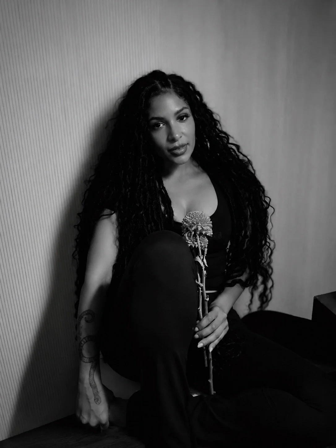 A woman with long, curly hair sitting on the floor, smiling and looking to the side, in a black dress with a tattoo on her left forearm, in front of a textured wall.