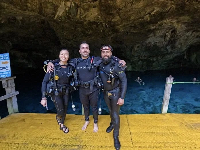 Three scuba divers standing on a yellow platform in a cave with scuba gear. There are people swimming in the blue water behind them.