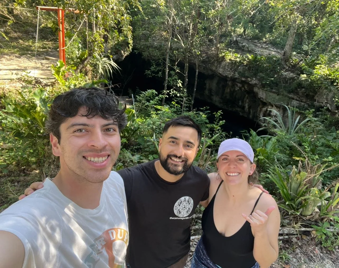 Three people taking a selfie in front of a cave entrance surrounded by lush greenery.