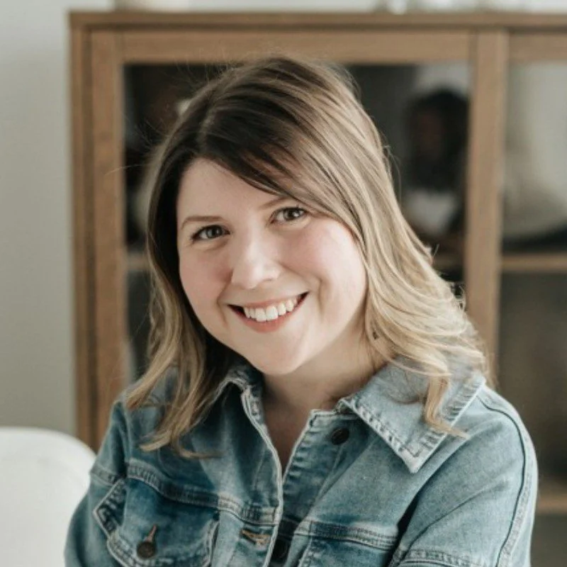 Une femme souriante avec des cheveux bruns mi-longs porte une veste en jean dans un intérieur domestique.