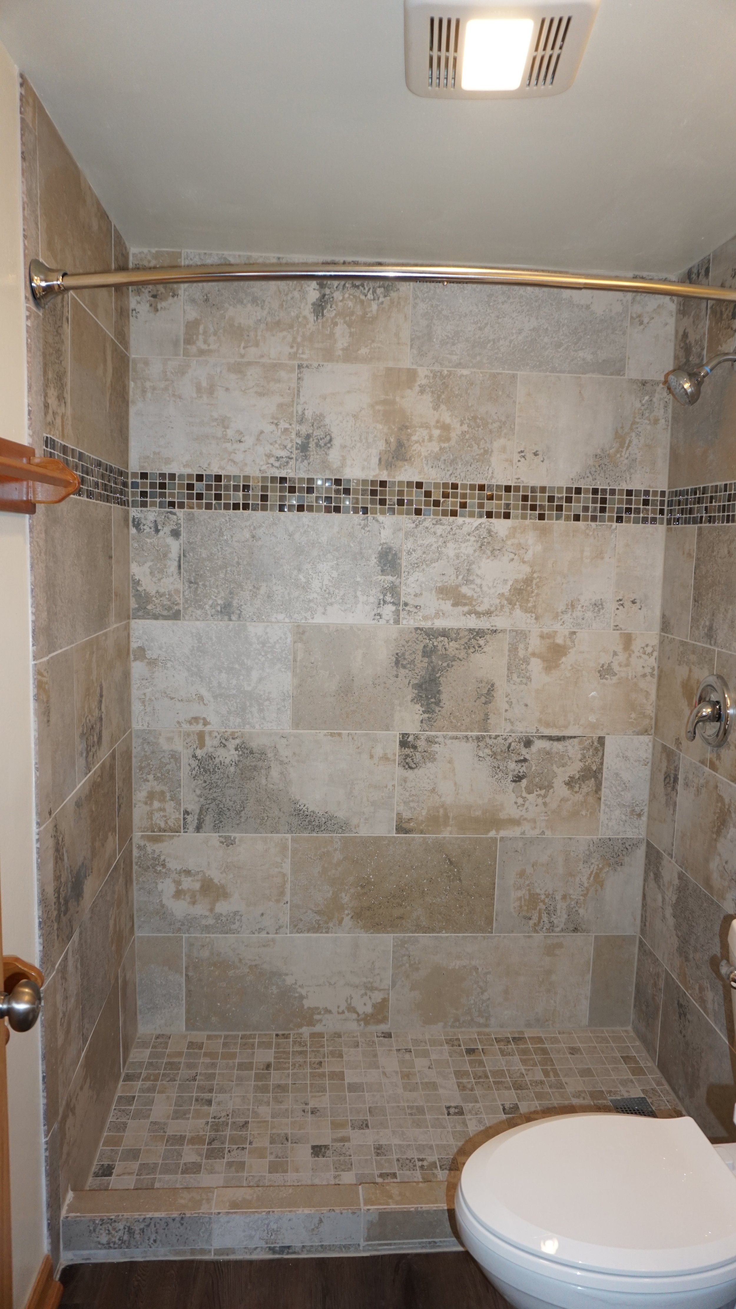 Bathroom with a shower area to the left, beige tiled walls, a white toilet in the center, a small dark cabinet with a white sink on top, a mirror above the sink, and a towel rack mounted on the wall.