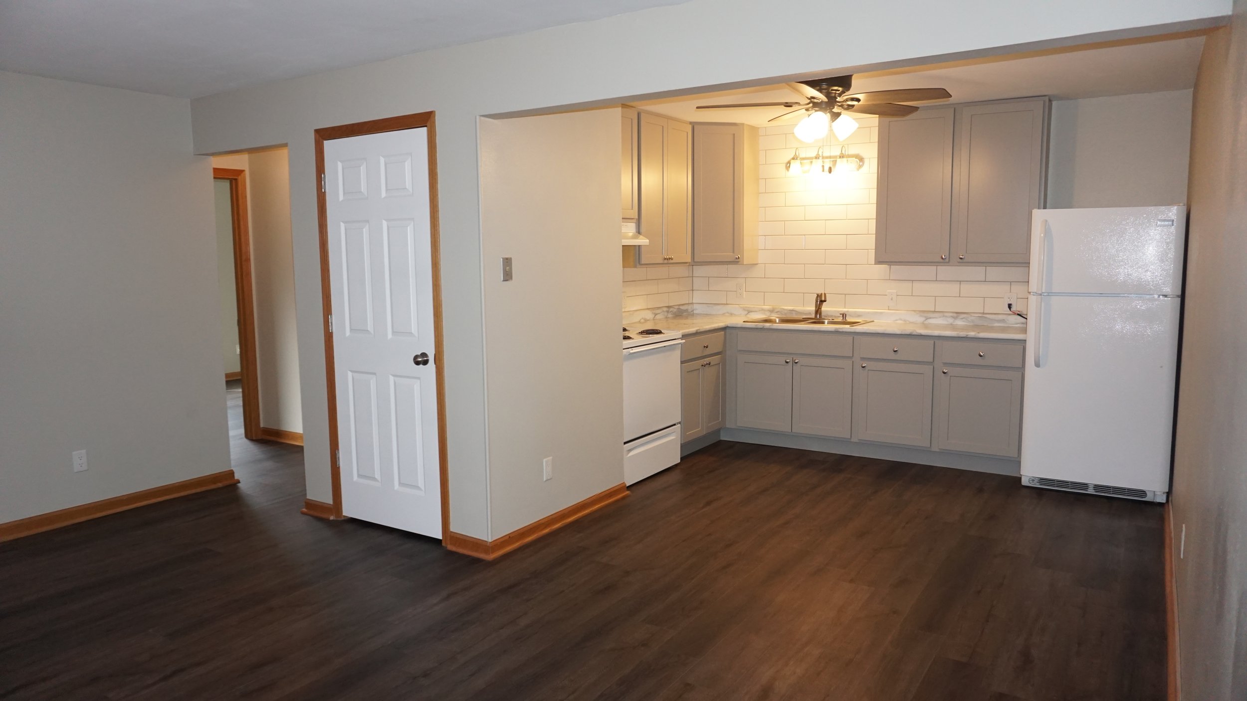 Empty living room and kitchen area with hardwood floors, white walls, and natural light coming through windows, showing shadows on the wall.