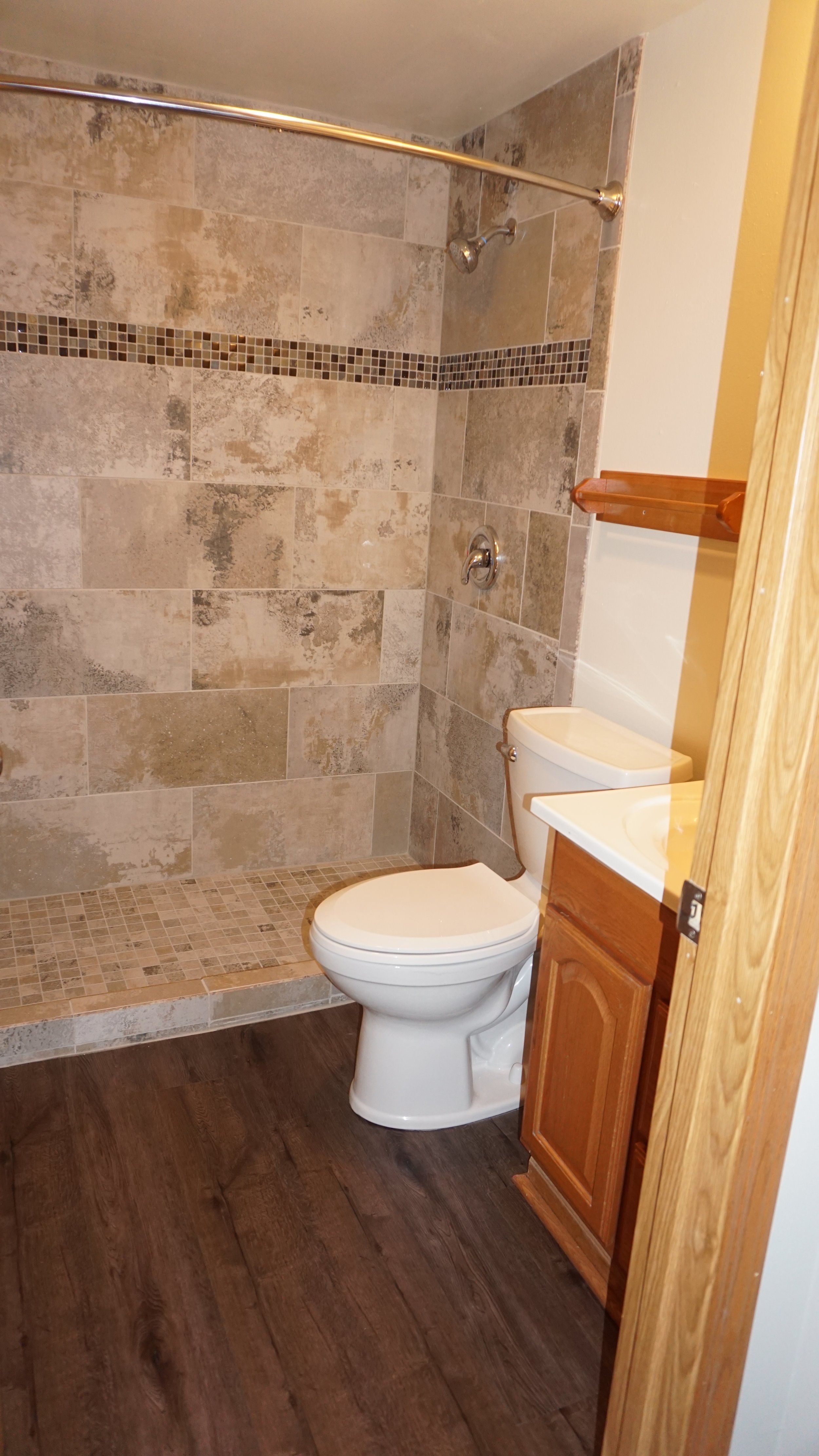 Bathroom with a shower area to the left, beige tiled walls, a white toilet in the center, a small dark cabinet with a white sink on top, a mirror above the sink, and a towel rack mounted on the wall.