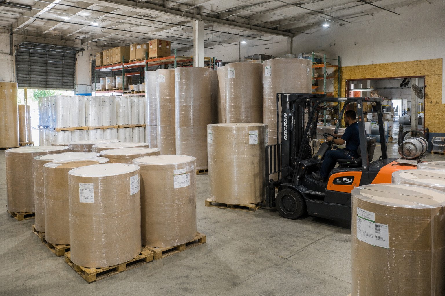 A warehouse with large paper rolls stored on pallets, and a forklift moving inventory.