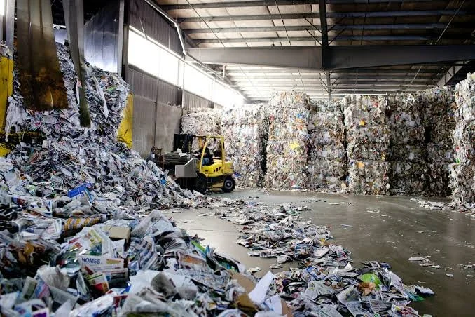 Large indoor recycling facility with towering stacks of compressed paper and cardboard, scattered loose paper on the floor, and a yellow forklift in the background.