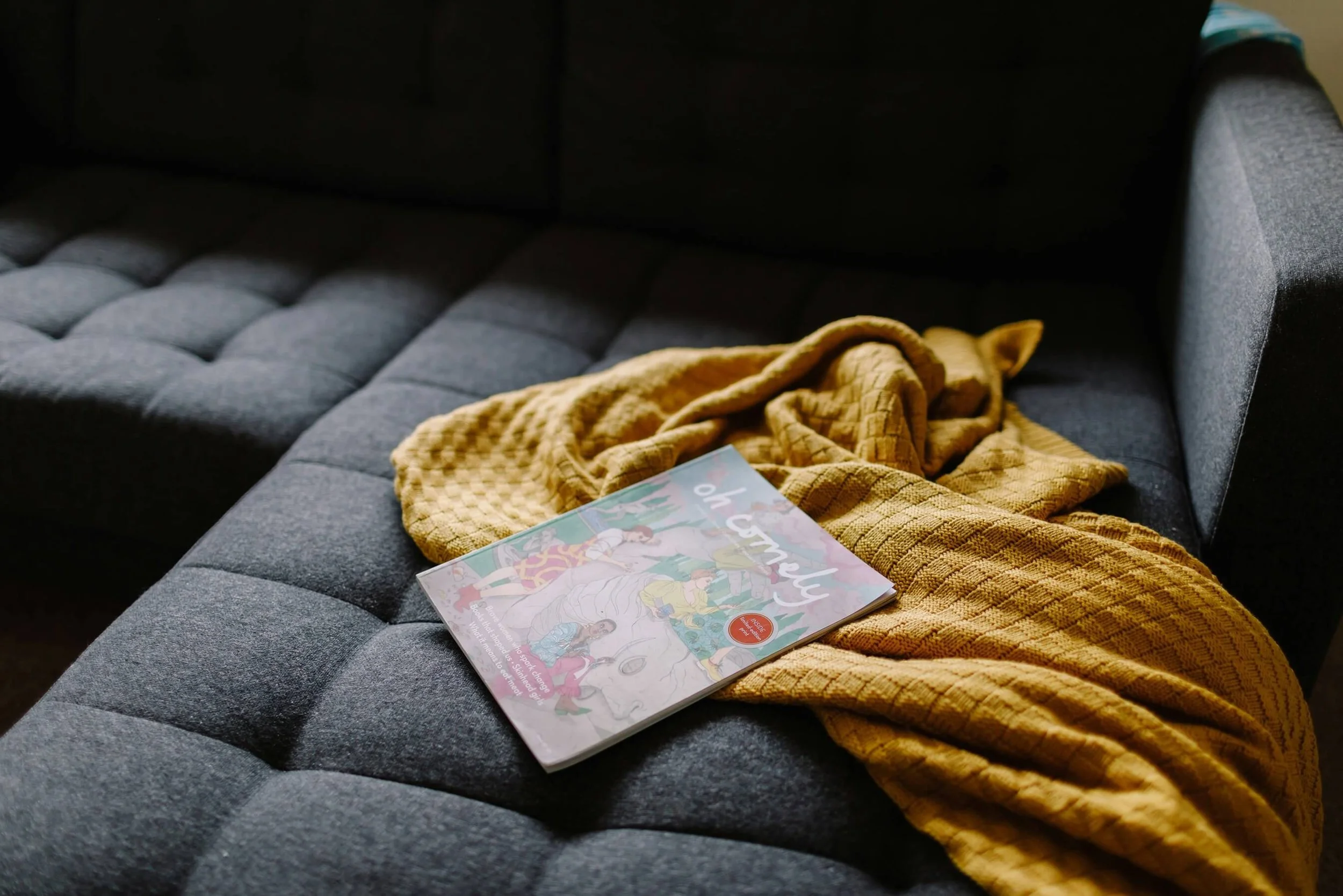 A gray upholstered couch with a yellow textured blanket and a colorful illustrated magazine on it.