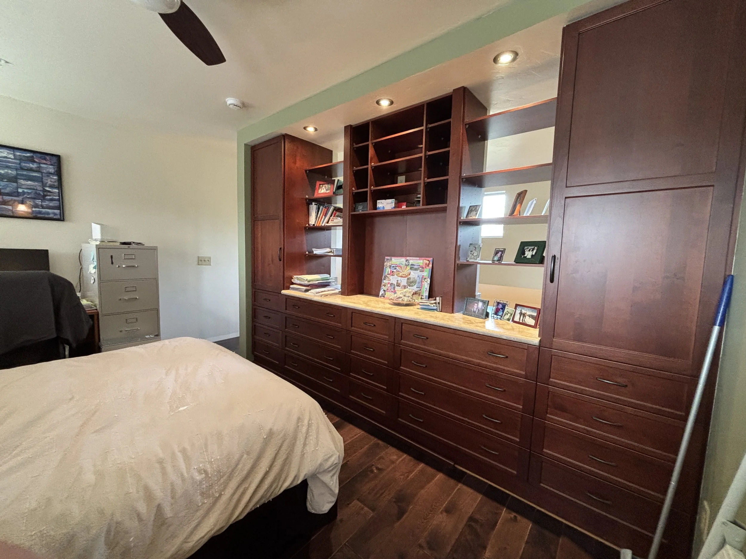 View of a bedroom corner featuring a bed with cream-colored quilt, a metal filing cabinet, a wooden wall unit with shelves and drawers, framed photographs, and personal items on the shelves.
