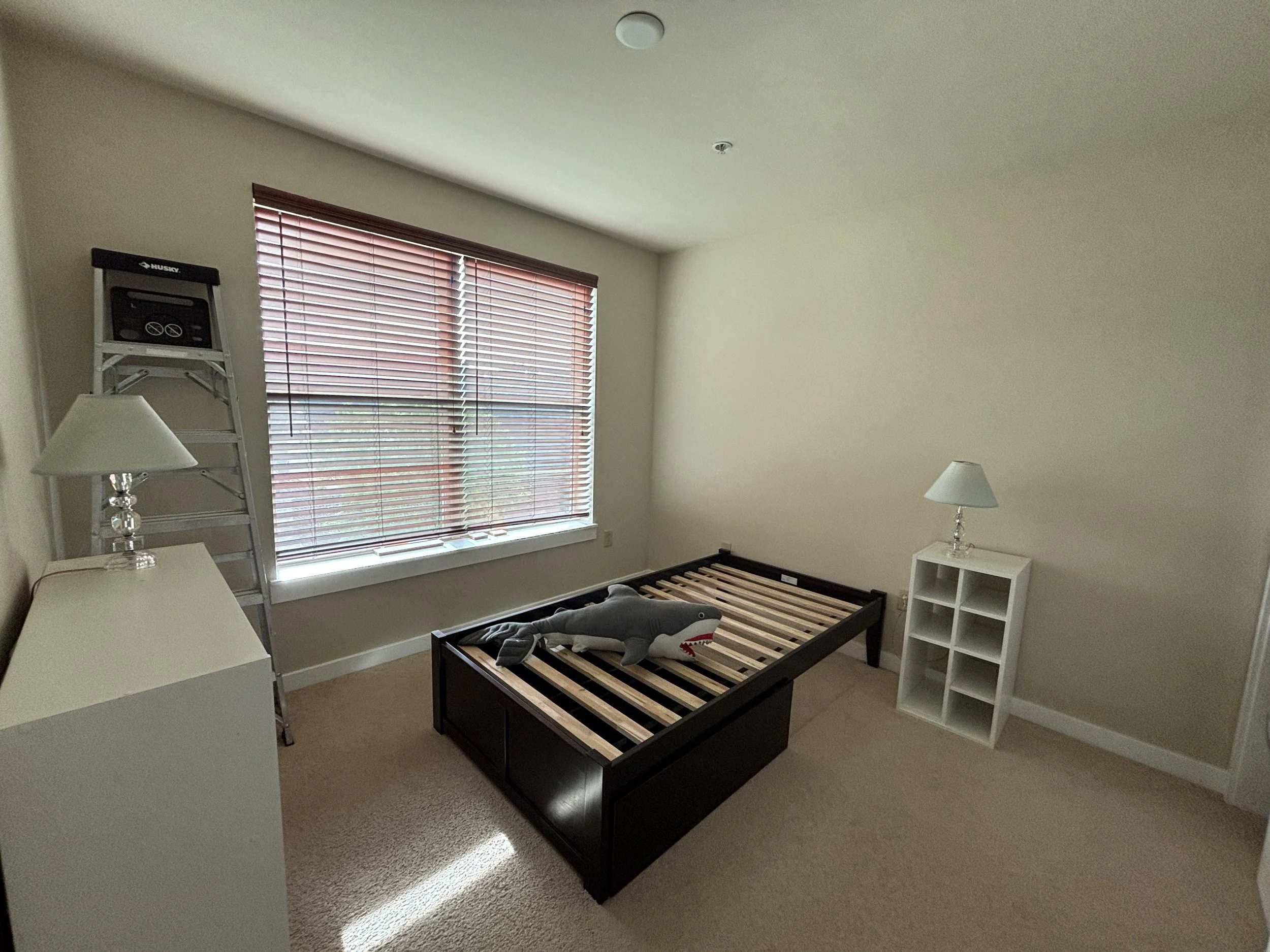 Empty bedroom with a bed frame, two white side tables with lamps, a shelf with a folded shirt, and a window with closed blinds.