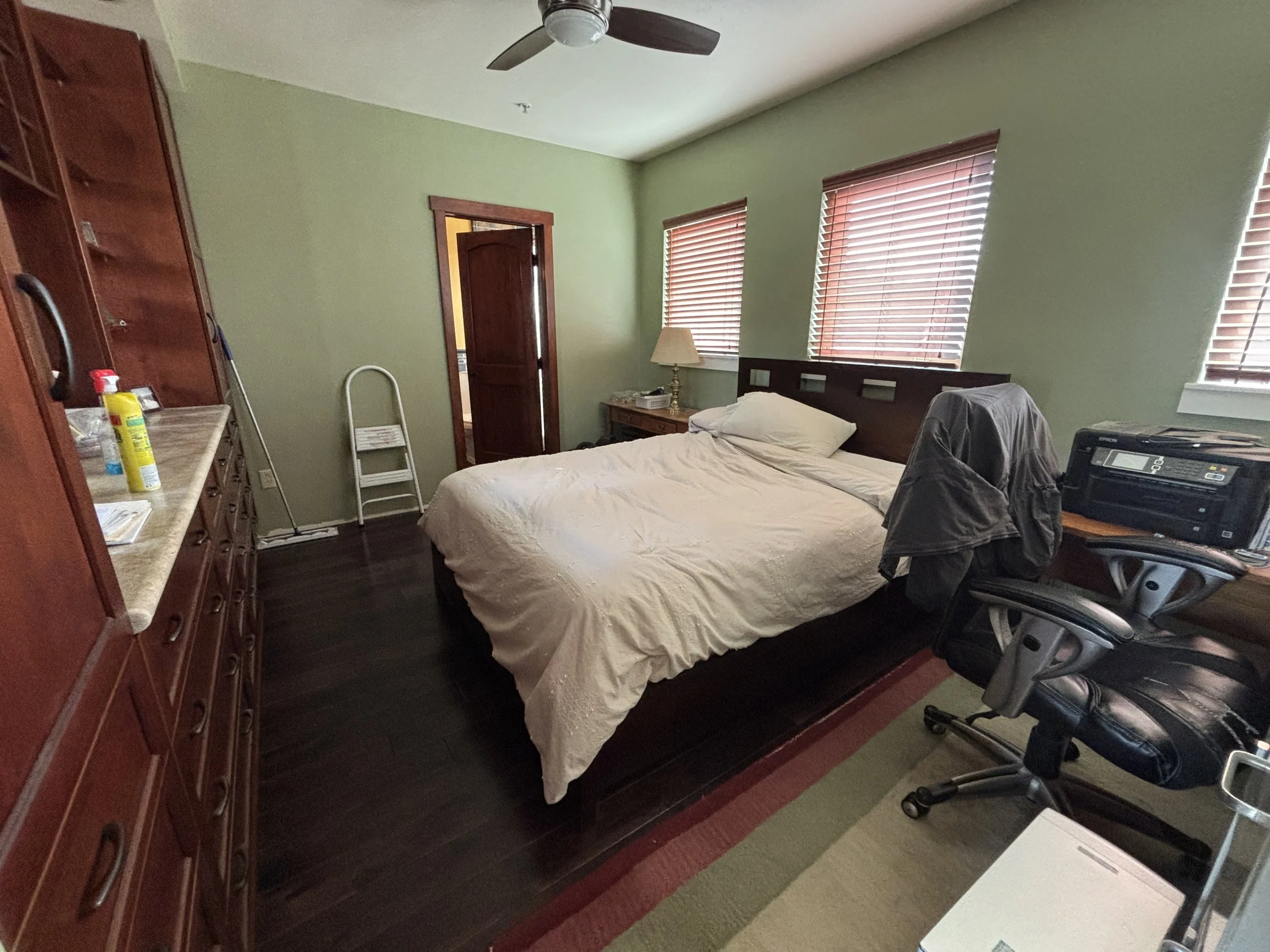 A bedroom with light green walls, wooden trim, and three windows with wooden blinds. There's a bed with white sheets, a nightstand with a lamp, a black office chair, a desk phone, and a dresser with various items on it. A small step stool and vacuum 