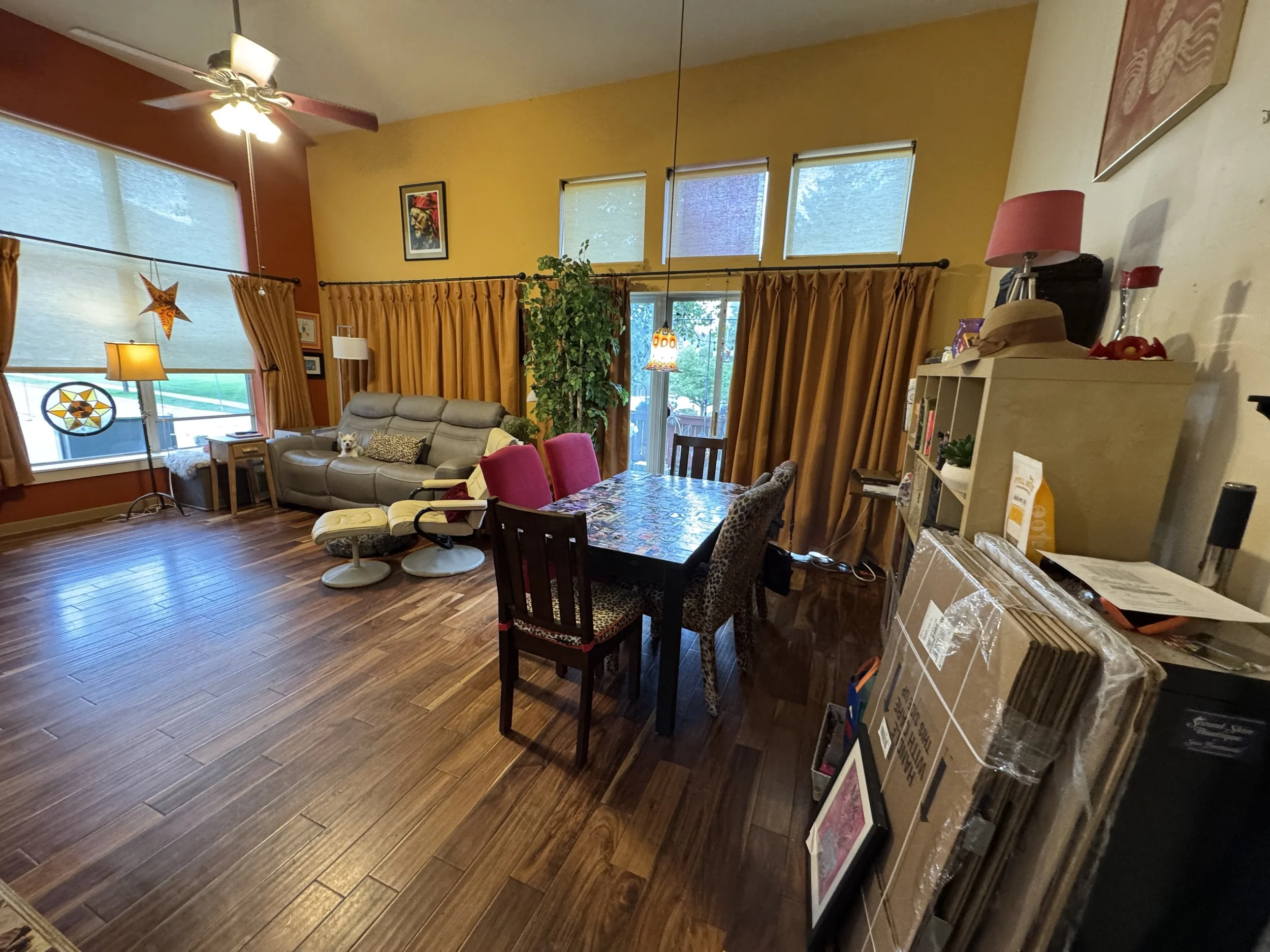 Living room with large windows, yellow and red walls, a ceiling fan, a gray couch, a wooden dining table with pink and leopard-print chairs, and a white bookshelf filled with books and decorative items. There are also plants, lamps, and art on the wa