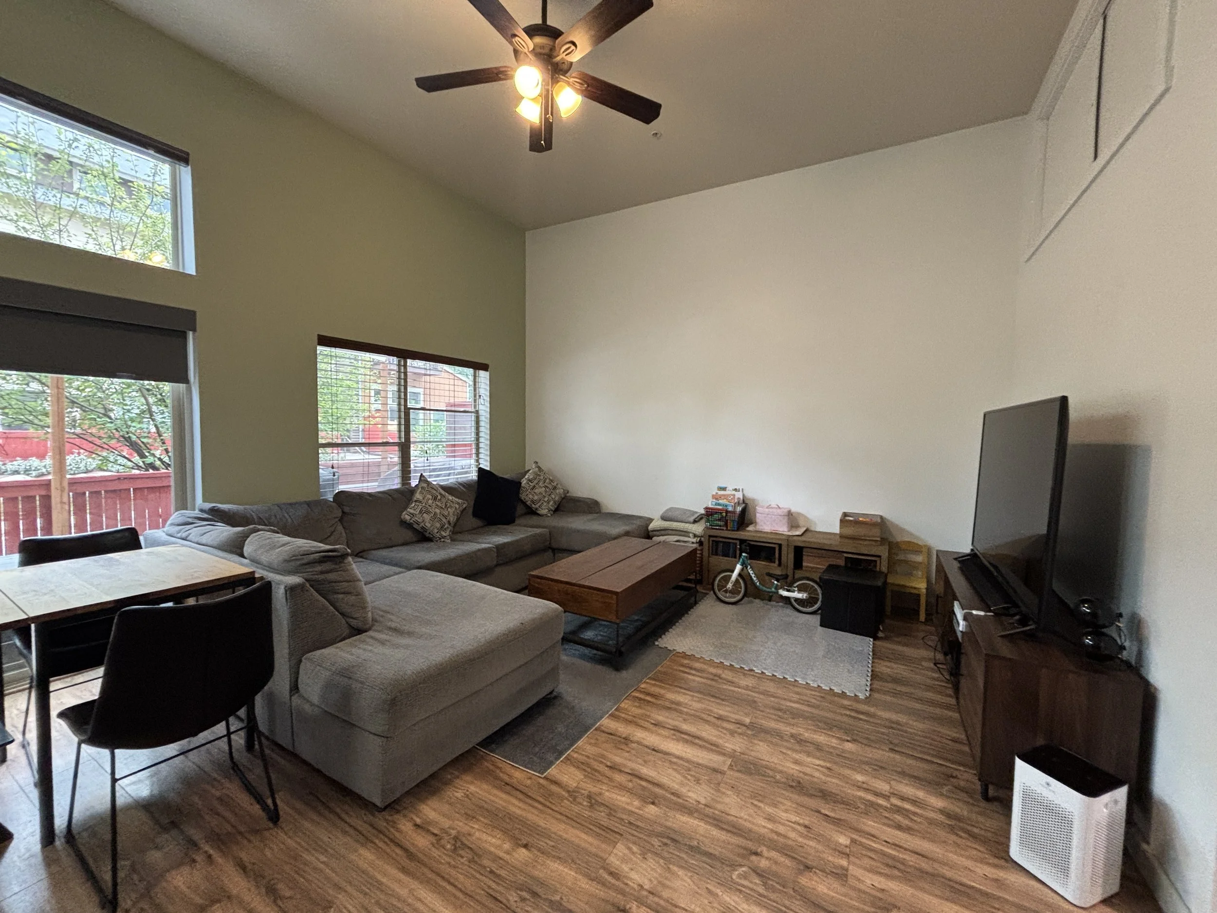 Living room with beige sectional sofa, black chairs, wooden coffee table, TV, large window with blinds, and ceiling fan with lights.