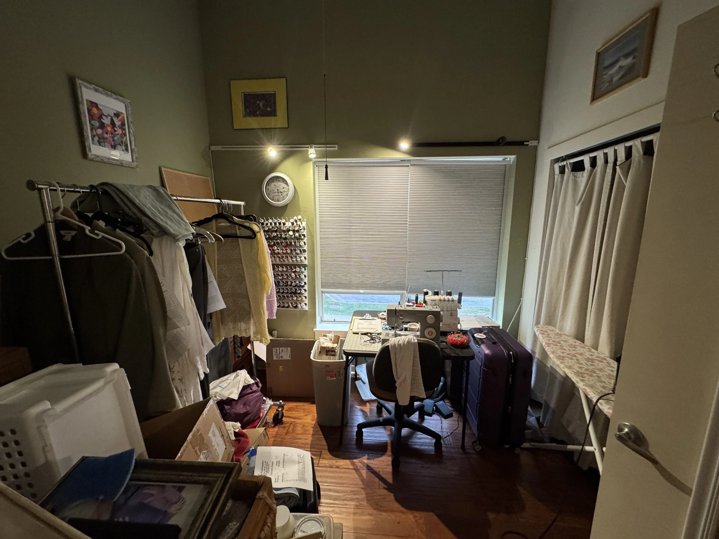 A cluttered room with various items including clothes hanging on a rack, a sewing machine, boxes, and framed pictures on the walls, with closed blinds covering a window.