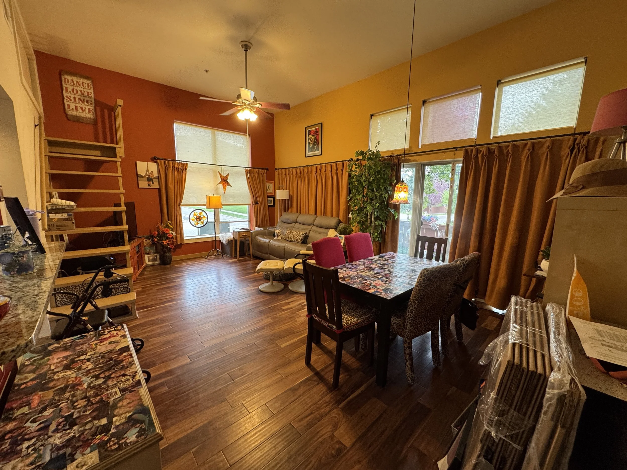 A cozy living room with yellow and red painted walls, large windows with beige curtains, and a ceiling fan with light. There is a beige sofa, a dining table with colorful tablecloth, and various decorative items including framed pictures, a potted pl