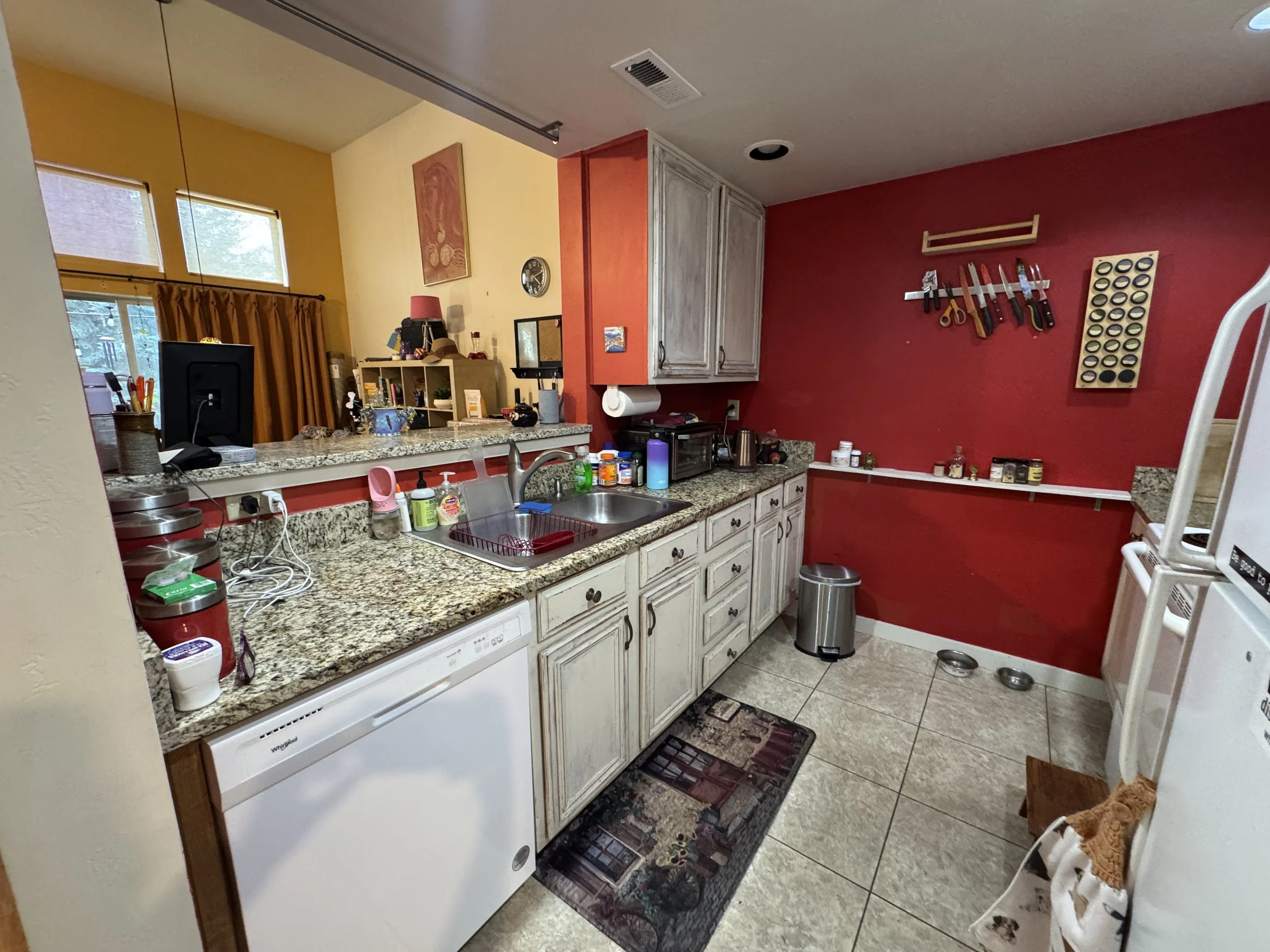 Kitchen with yellow and red painted walls, granite countertops, white cabinets, and various kitchen appliances and utensils. There is a window with yellow curtains, a dish drying rack, and a coffee station. Kitchen knives are hanging on the red wall.