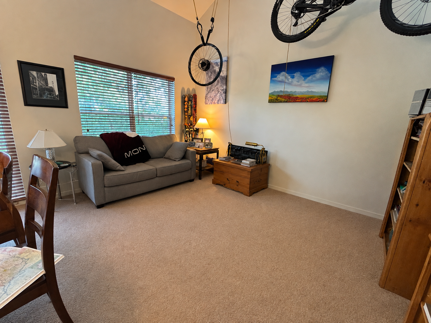 A cozy living room with a beige carpet, gray sofa with pillows and a blanket, a side table with a lamp, ski equipment against the wall, and wall art of a mountain scene. There's a wooden chest with books, a rack with bicycles mounted upside down on t