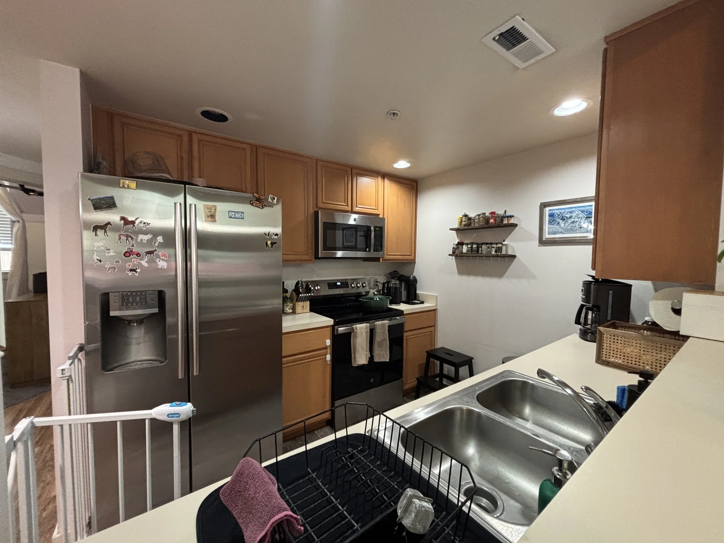 A kitchen with wooden cabinets, stainless steel refrigerator with dog magnets, microwave, stove, and a countertop with a sink. There are various kitchen items and a small frame picture on the wall, a small black stool, and a child gate in the foregro