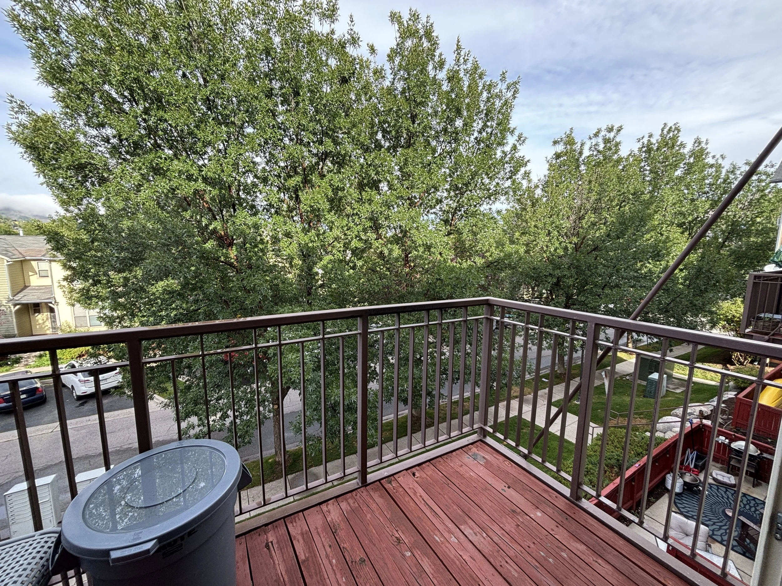 A view from a balcony shows a wooden deck with a round plastic outdoor table and chairs, a large tree with dense green leaves in the background, parked cars on the street, and parts of neighboring buildings under a partly cloudy sky.