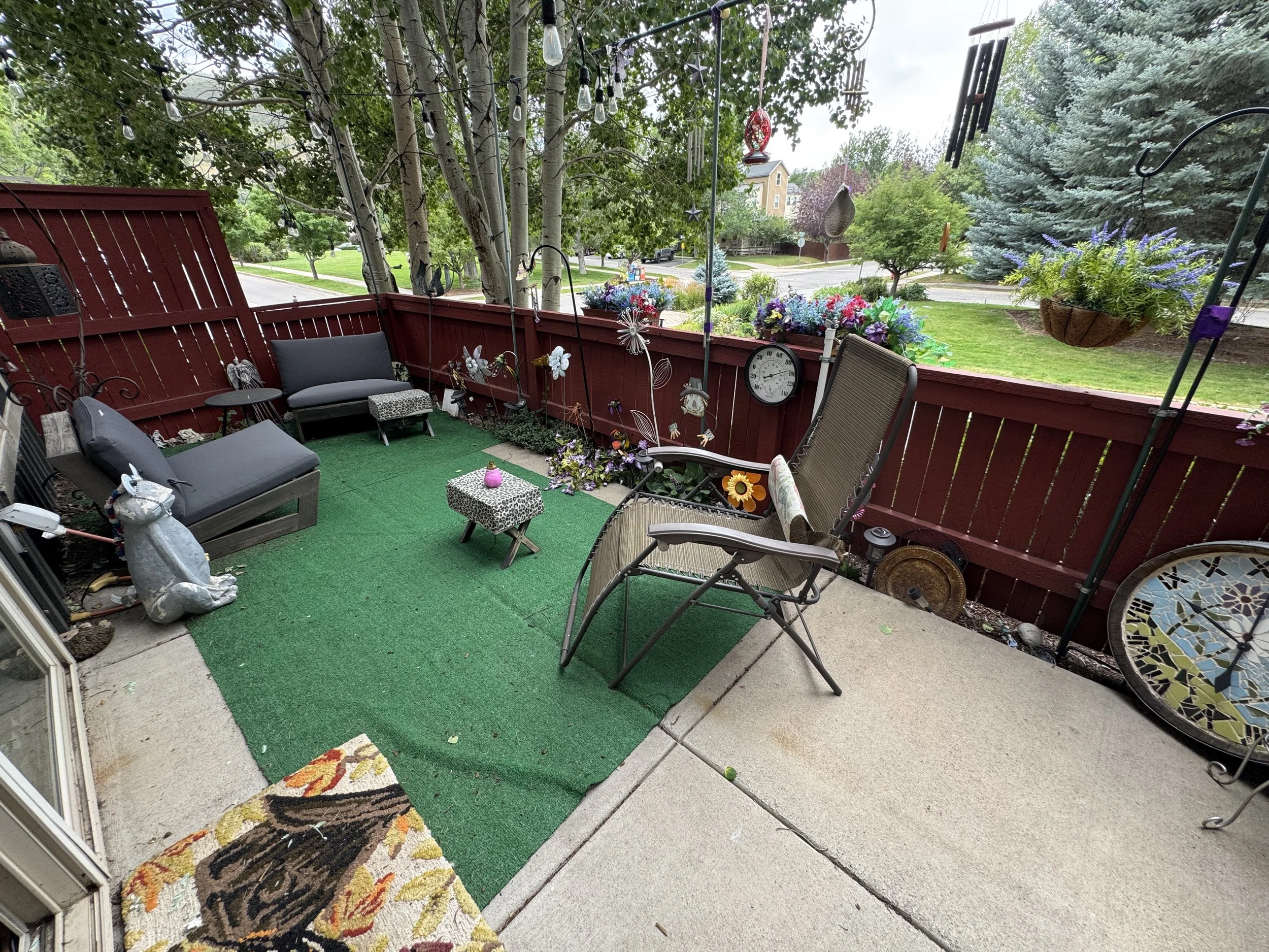 A decorated outdoor porch with patio furniture, hanging wind chimes, various planters with flowers, a garden gnome, a clock, and string lights, overlooking a green lawn and trees.